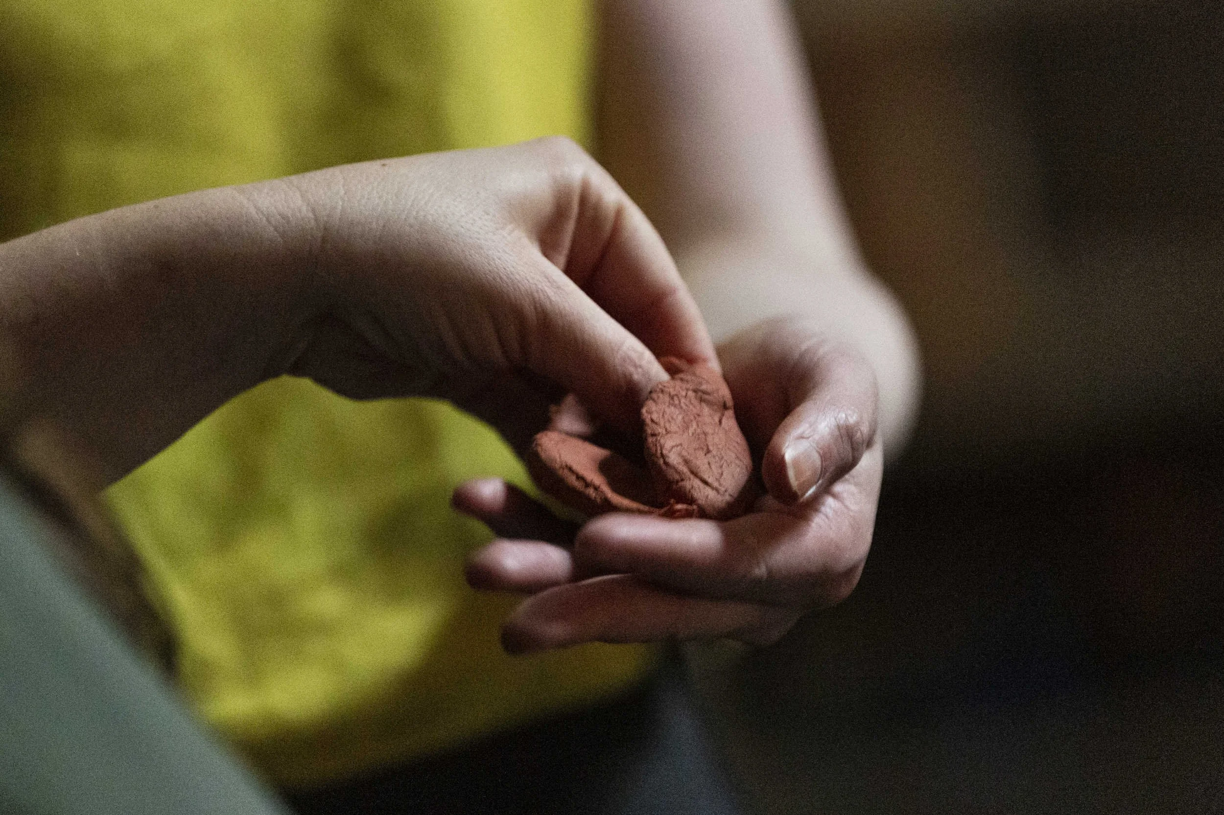Close-up of hands delicately holding and turning a small piece of terracotta clay, yellow fabric visible behind, in the Bodies of Water performance.
