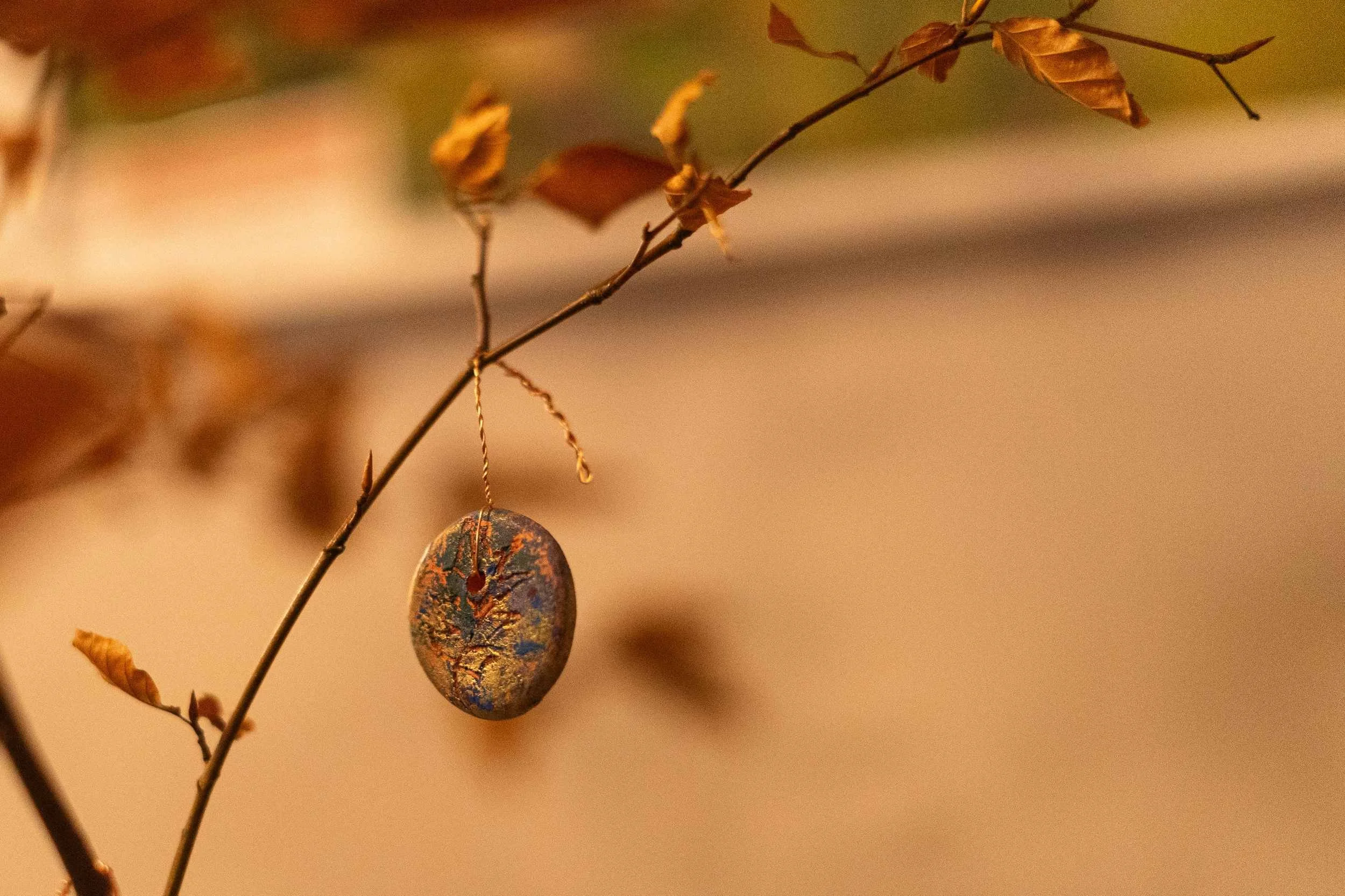 Decorated stone pendant hanging from an autumn branch with dried leaves, warm golden bokeh background.