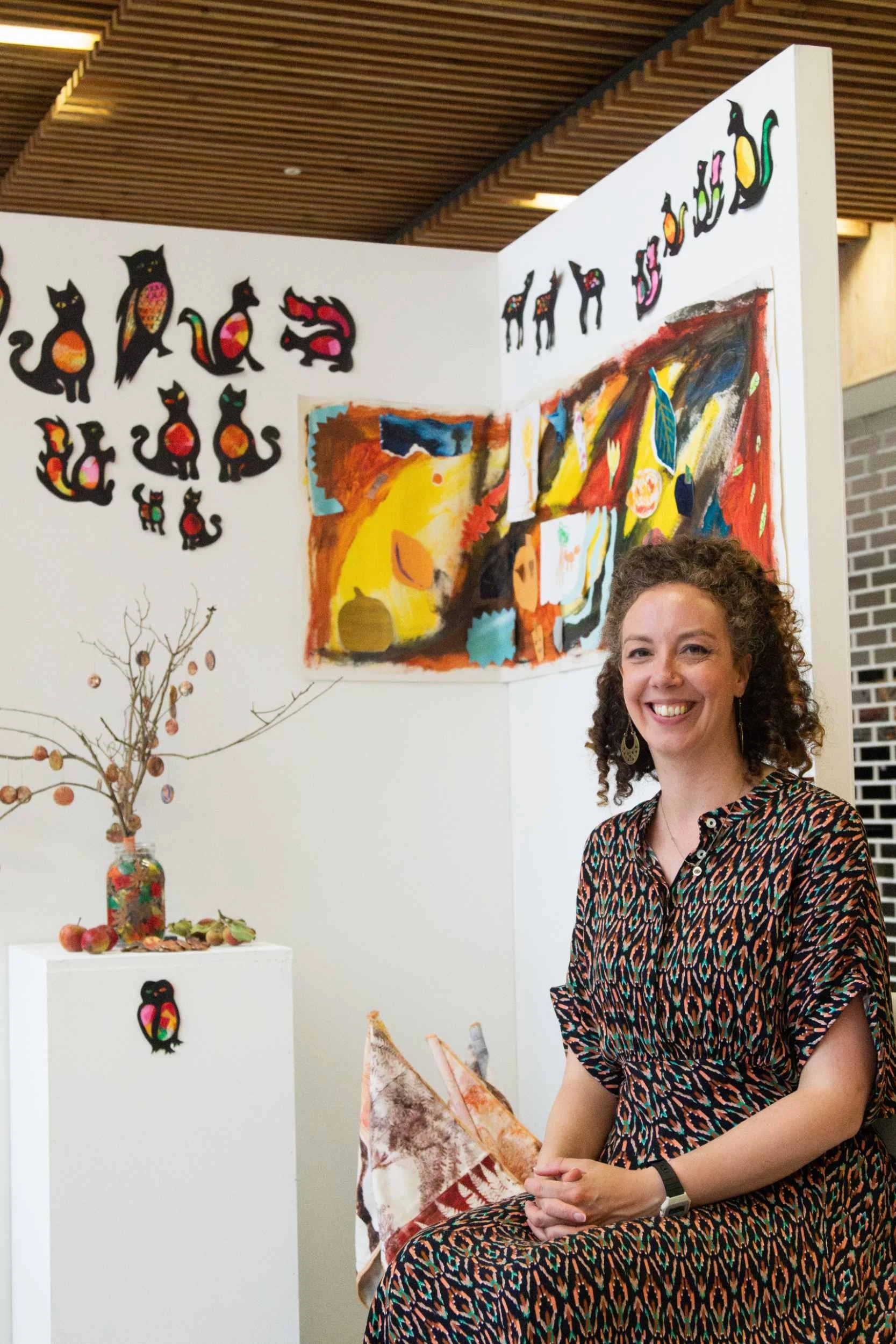 Woman smiles in front of a community art exhibition display featuring colourful animal cutouts and painted canvases in Remembering Together.