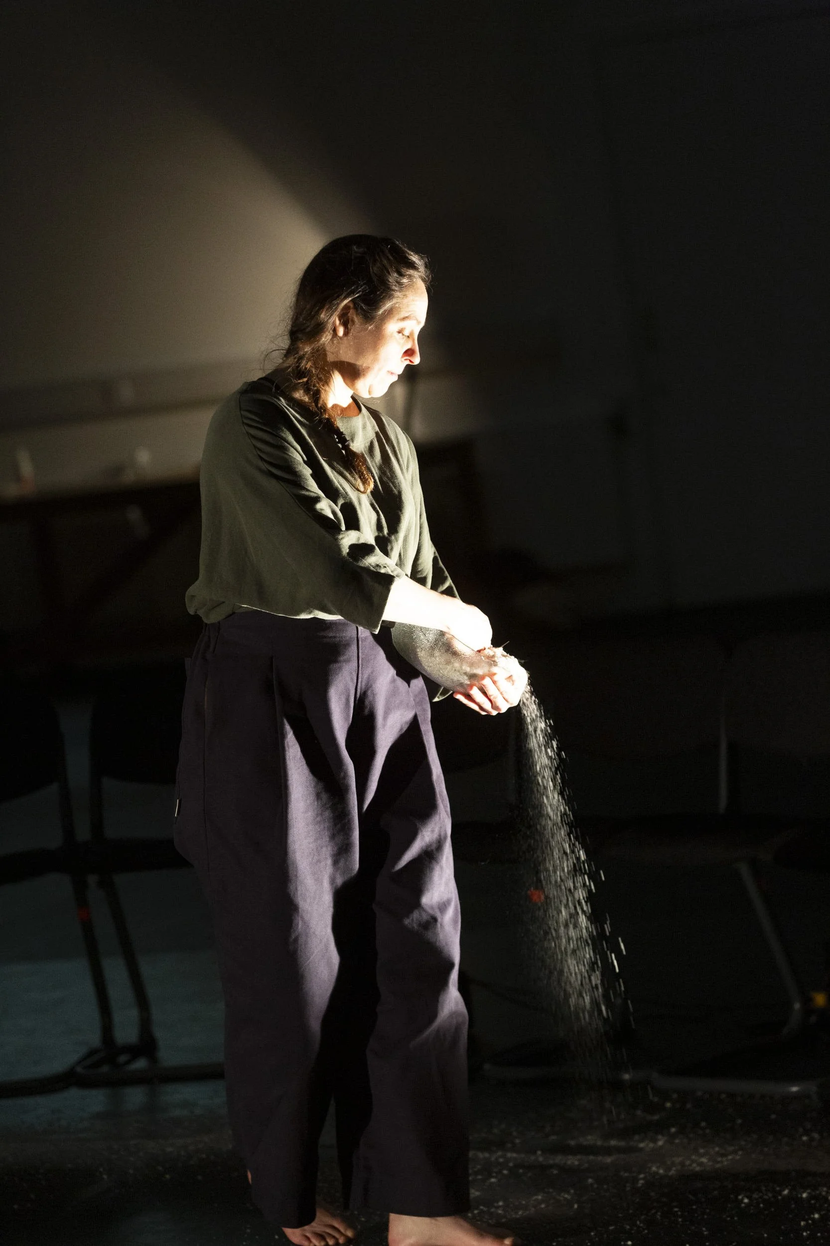 Performer in dark green stands in a strong shaft of light, releasing fine white powder that streams from her cupped hands in Bodies of Water.