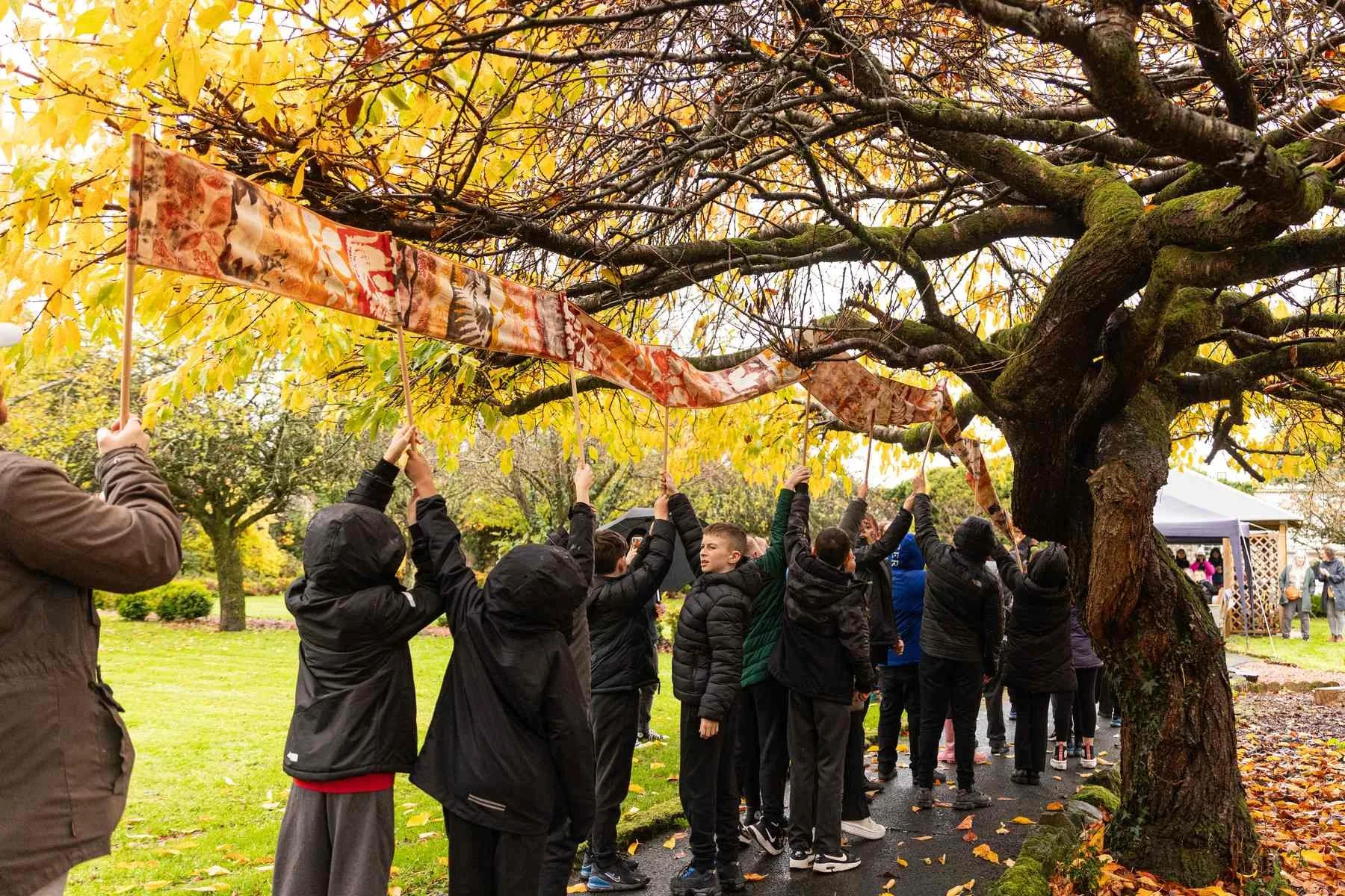 Line of children raise a long printed fabric banner overhead beneath golden autumn trees in a park on a rainy day.