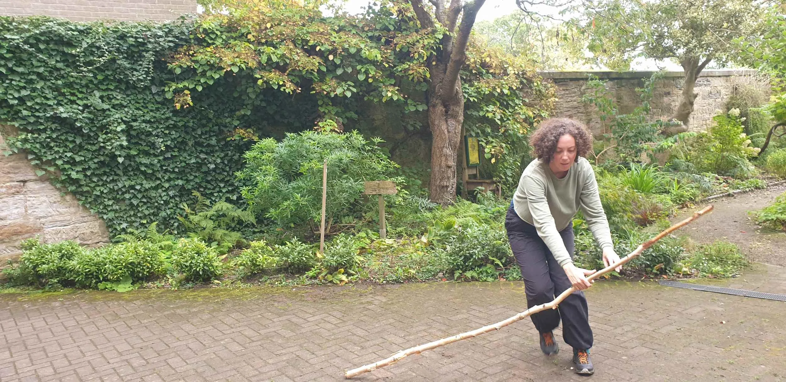 Dancer in sage green holds a long stick close to the ground in a walled garden with ivy-covered stone walls in the Garden Series.