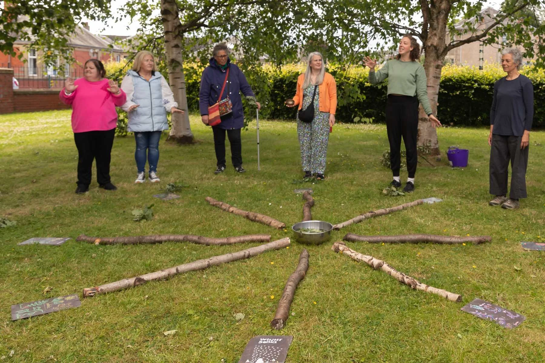 Six adults stand around a radial log installation on grass, led by a facilitator with arms open, trees behind.