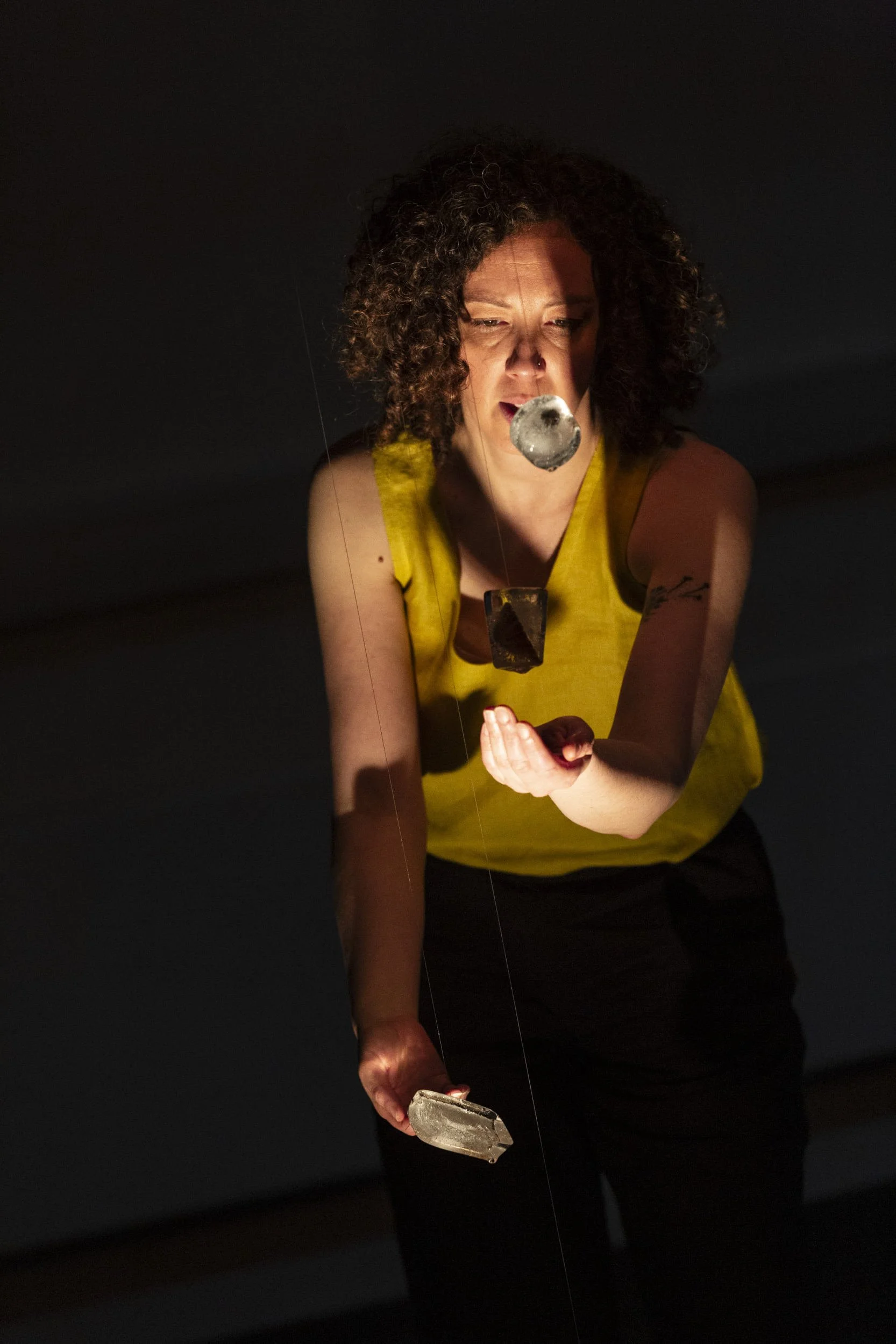 Performer in yellow leans forward in dramatic lighting, suspended ice block objects hanging before her, one hand extended in Bodies of Water.