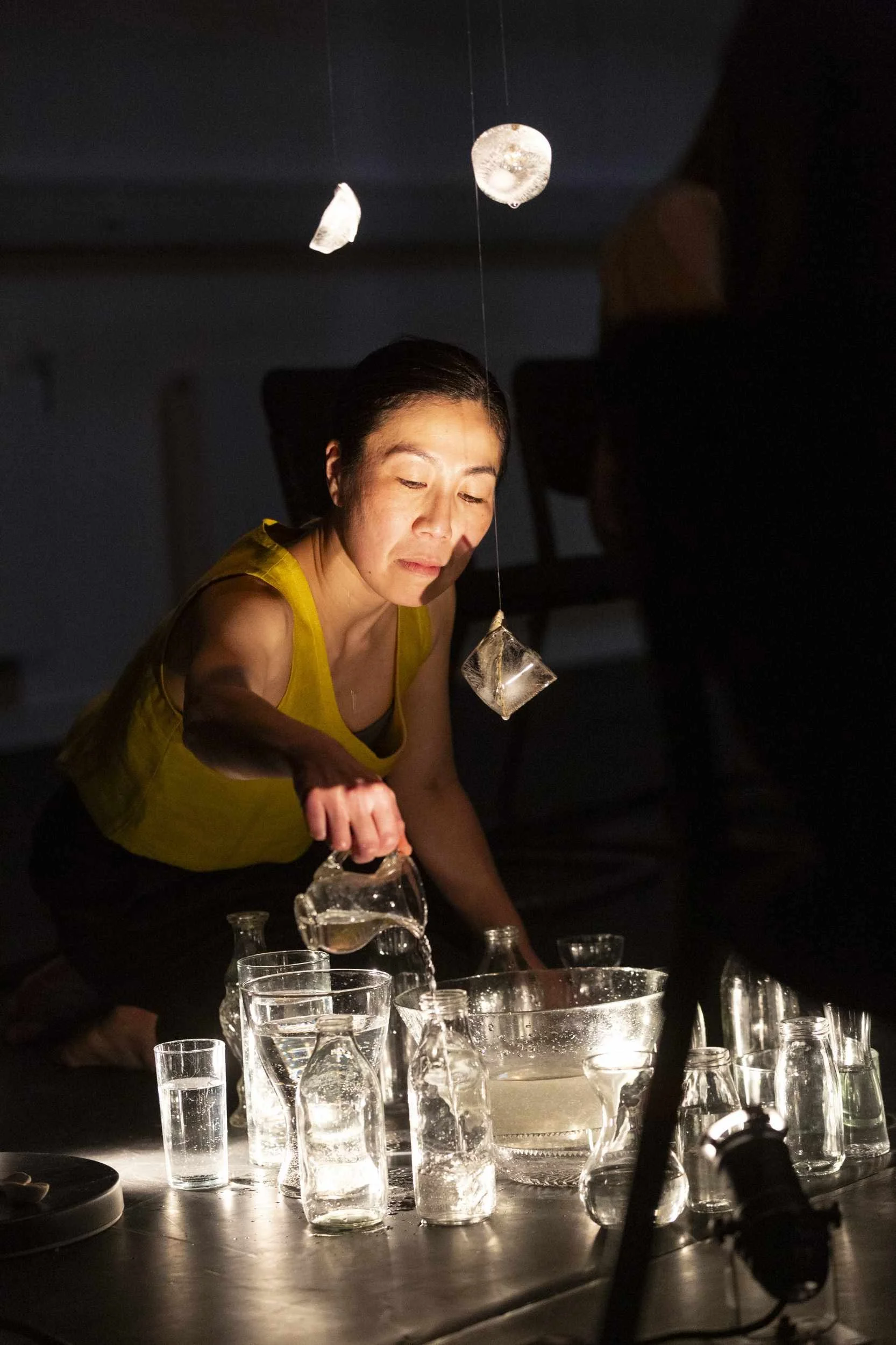 Performer in yellow leans over a table of illuminated glass vessels, pouring water from a jug, translucent objects suspended above in Bodies of Water.