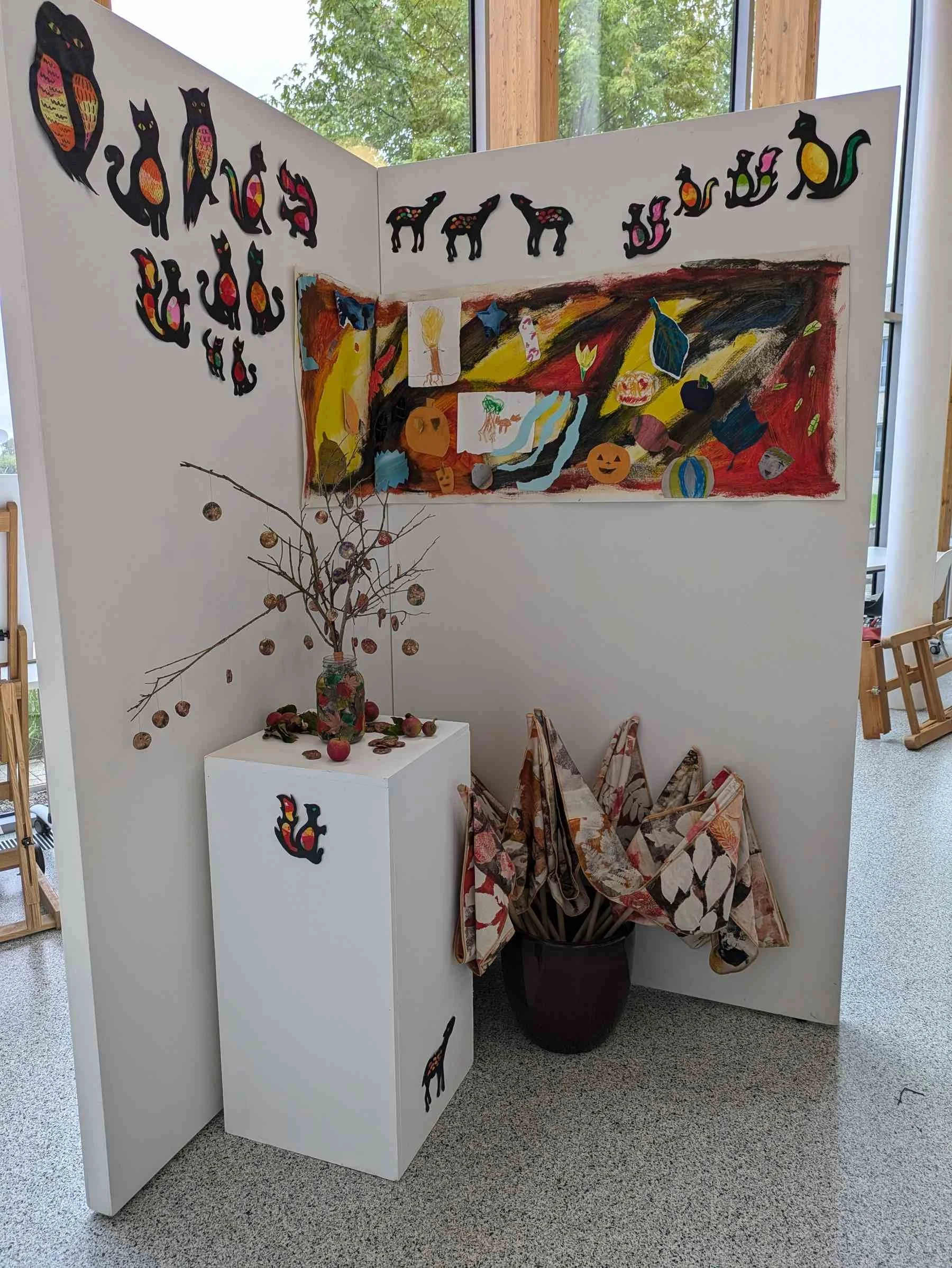 Exhibition display with cut-out animal silhouettes, a large collaborative autumn painting, branch installation, and printed fabric flags.