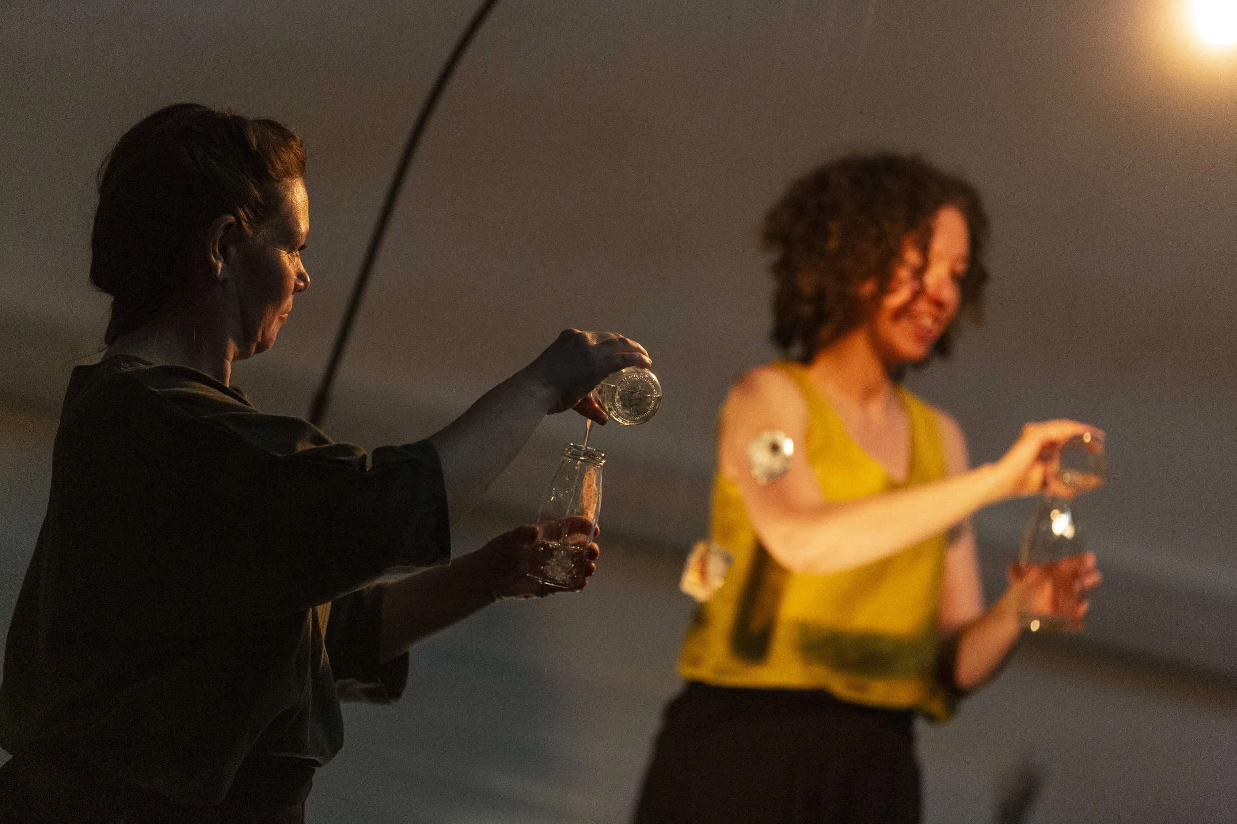 Performer in silhouette pours water between glass bottles in warm lamplight, a second performer in yellow visible behind in Bodies of Water.
