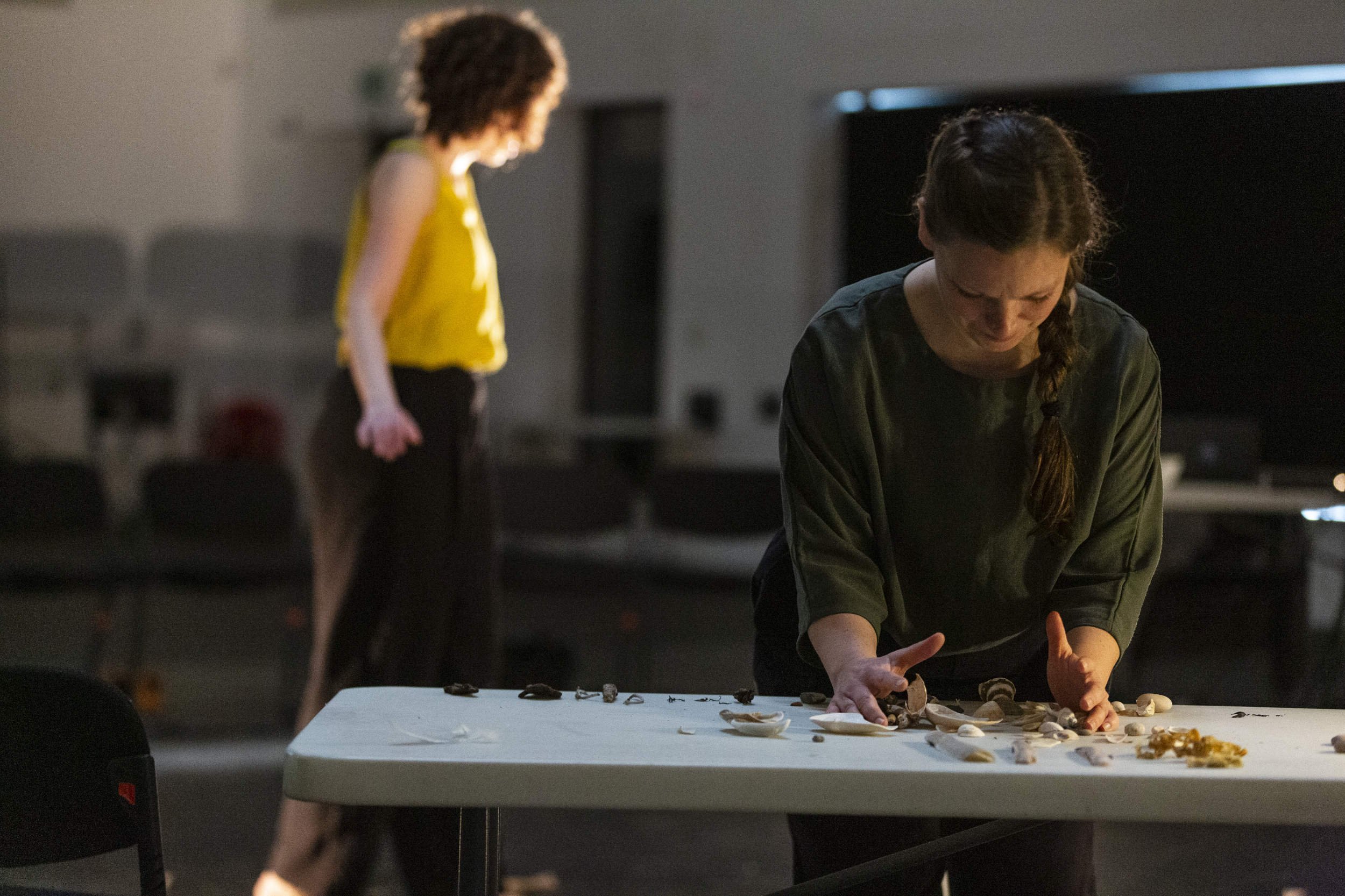 Performer in dark green works with shells and natural objects on a table, a second performer in yellow moving in soft focus behind in Bodies of Water.