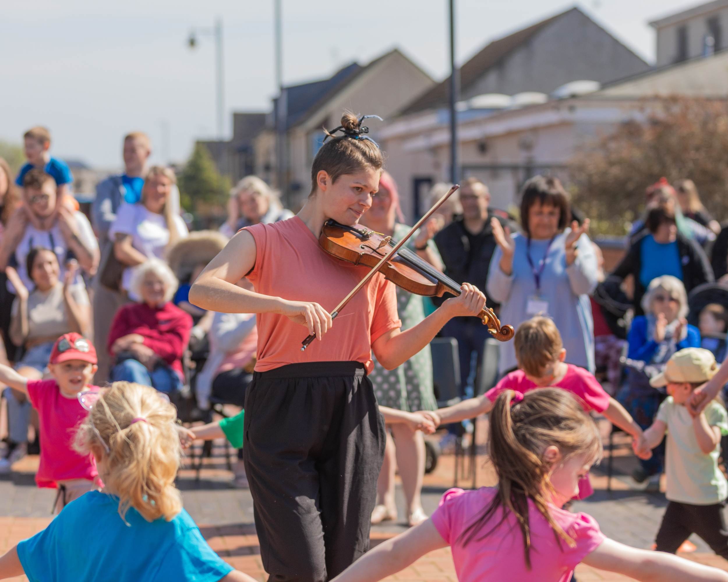 Children form a large circle dance on a sunny town square while a violinist in orange plays at the centre.