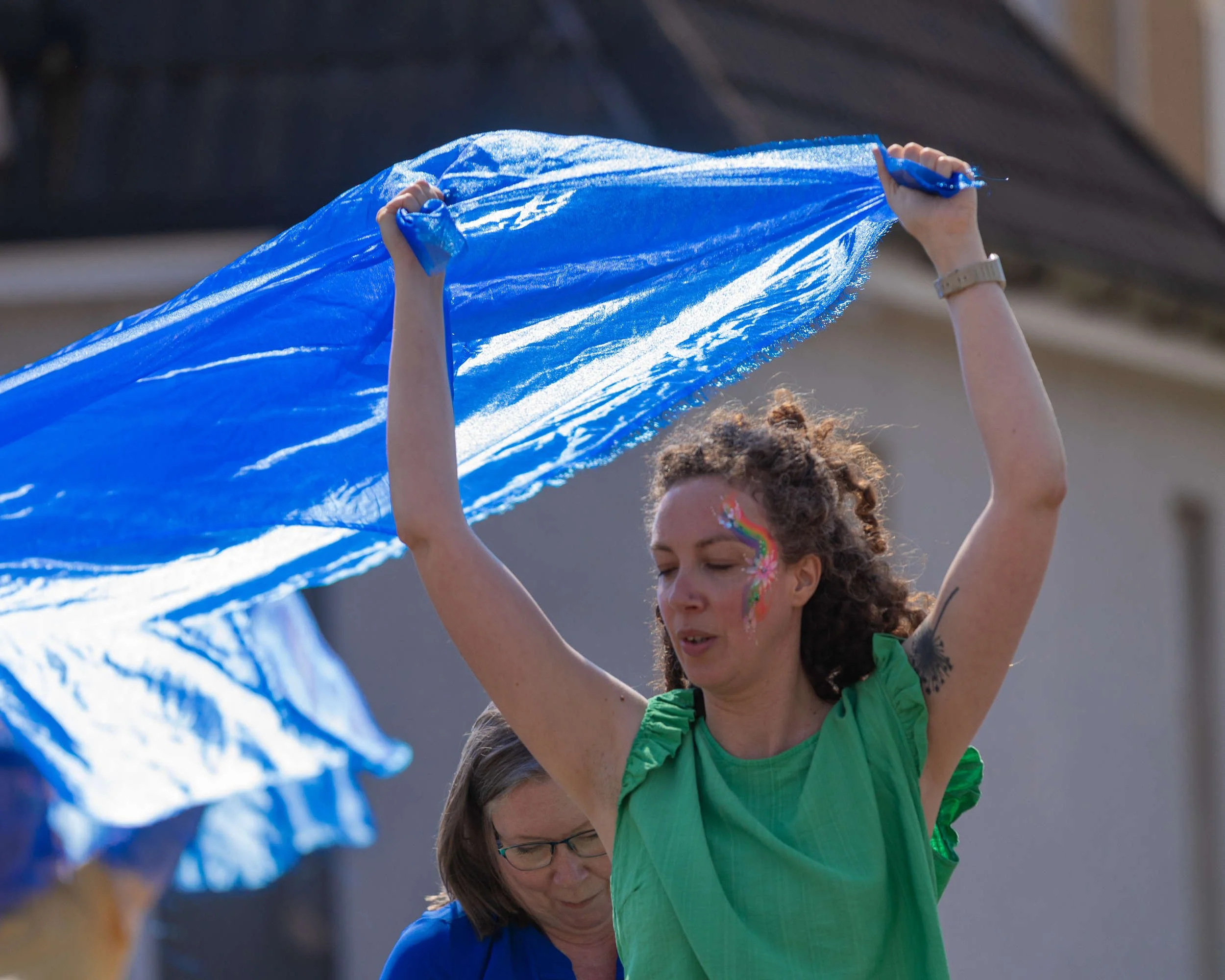 Performer in a green top with rainbow face paint raises a large shimmering blue fabric overhead during an outdoor Remembering Together performance.
