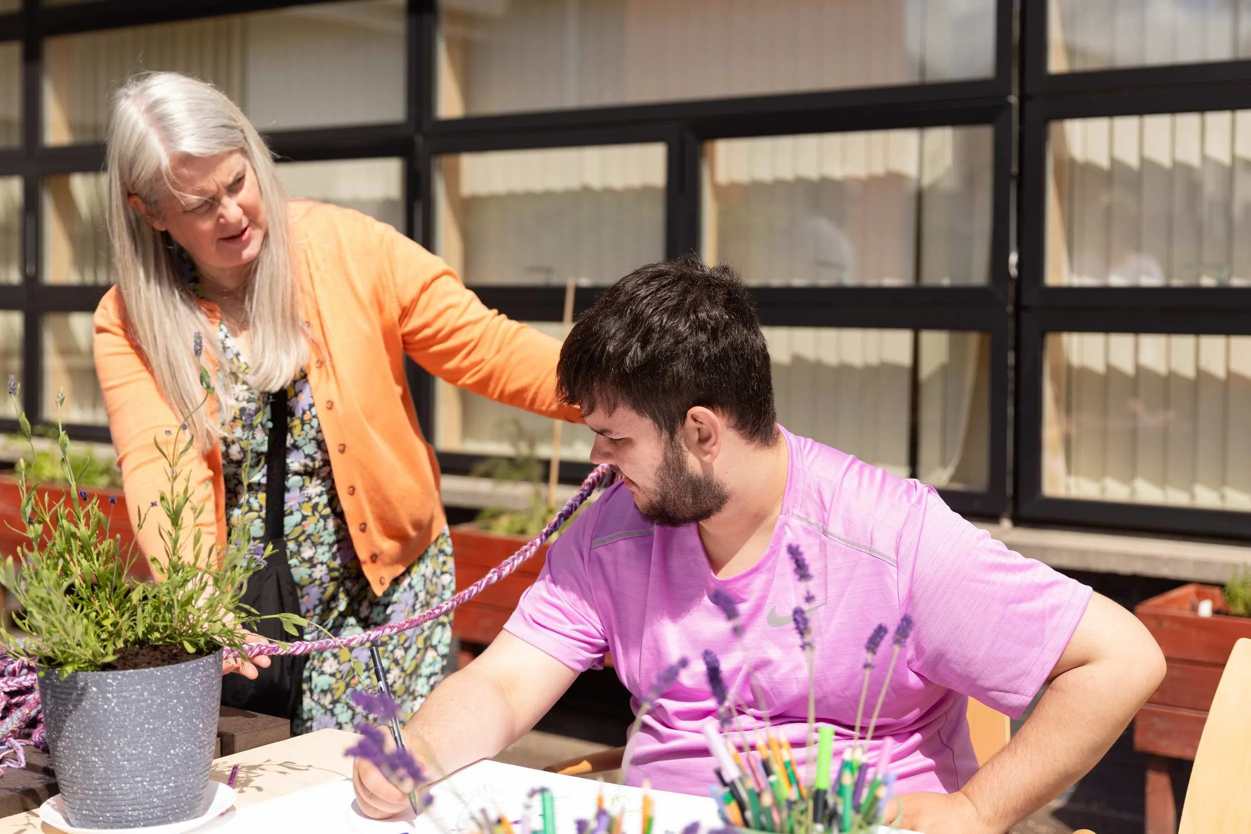 Older adult facilitator leans in to support a younger participant drawing at an outdoor table surrounded by lavender plants.