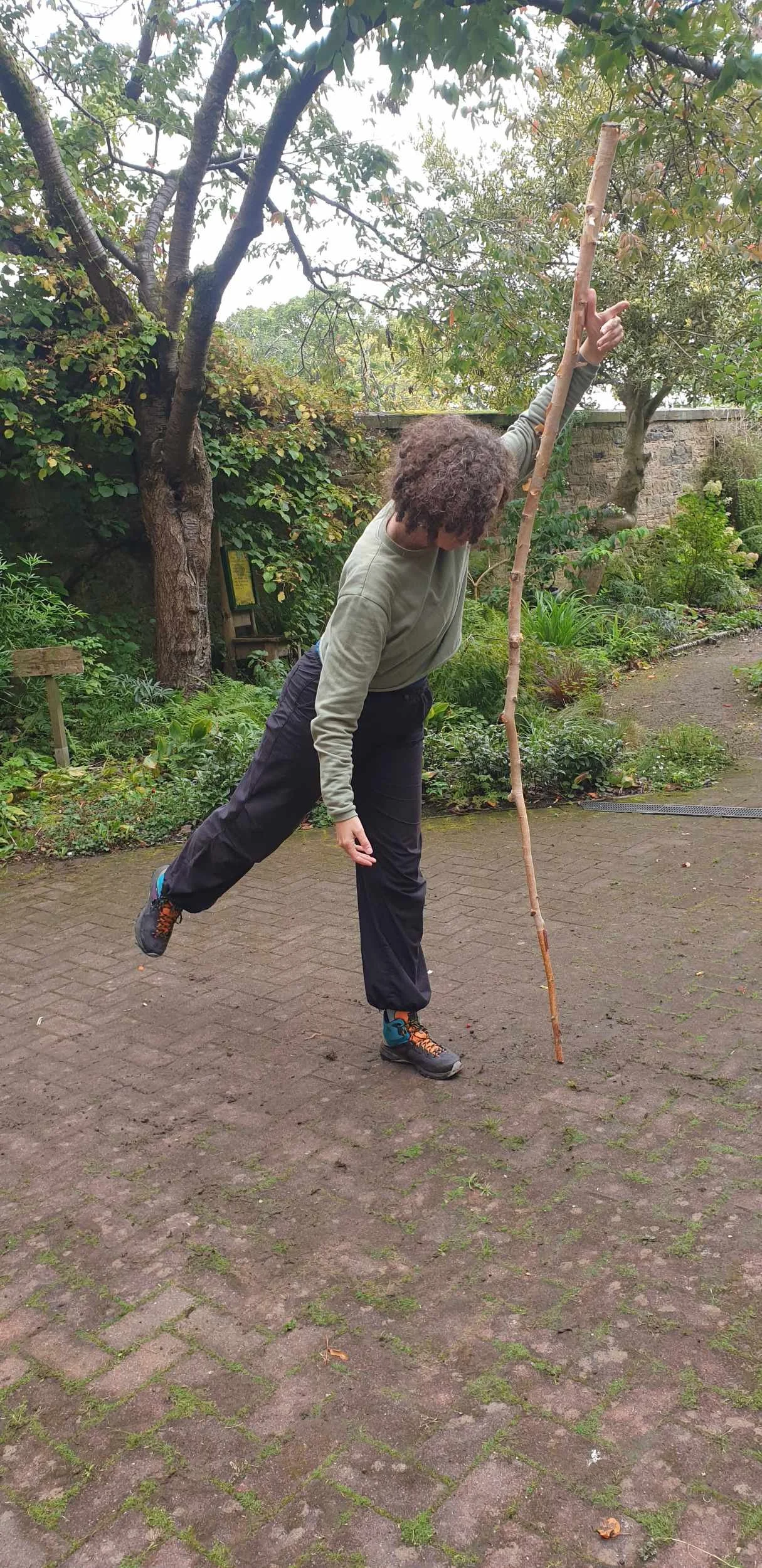 Dancer balances on one leg while holding a tall stick upright in a walled garden during a Garden Series session.