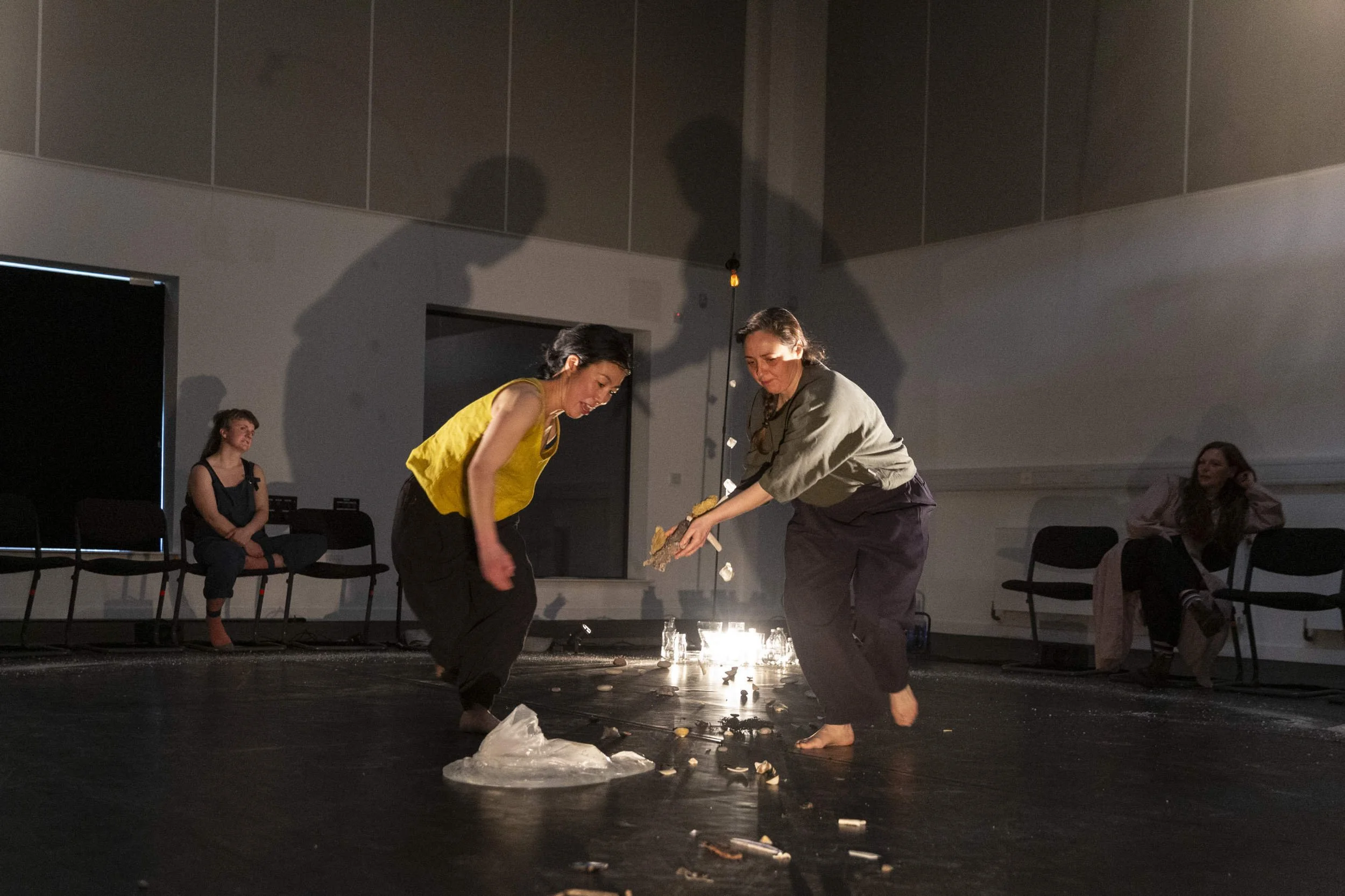 Two performers face each other over a glowing arrangement of glass bottles on a dark floor, audience members seated around them in Bodies of Water.