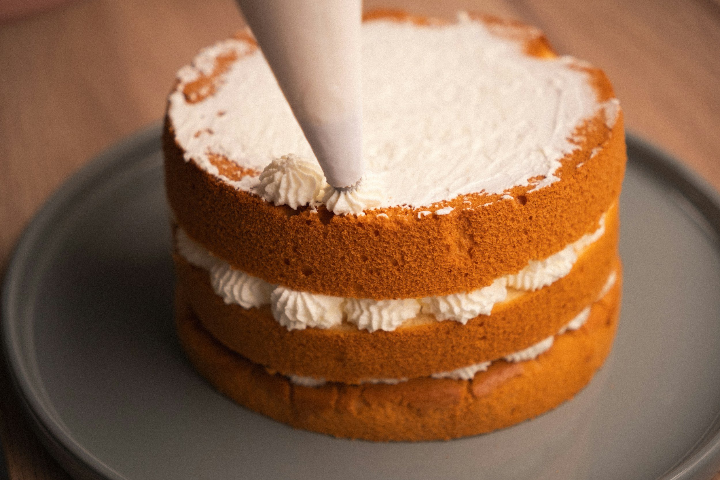 Three layered cake with whipped cream filling and a white frosting top, being decorated with a piping bag.