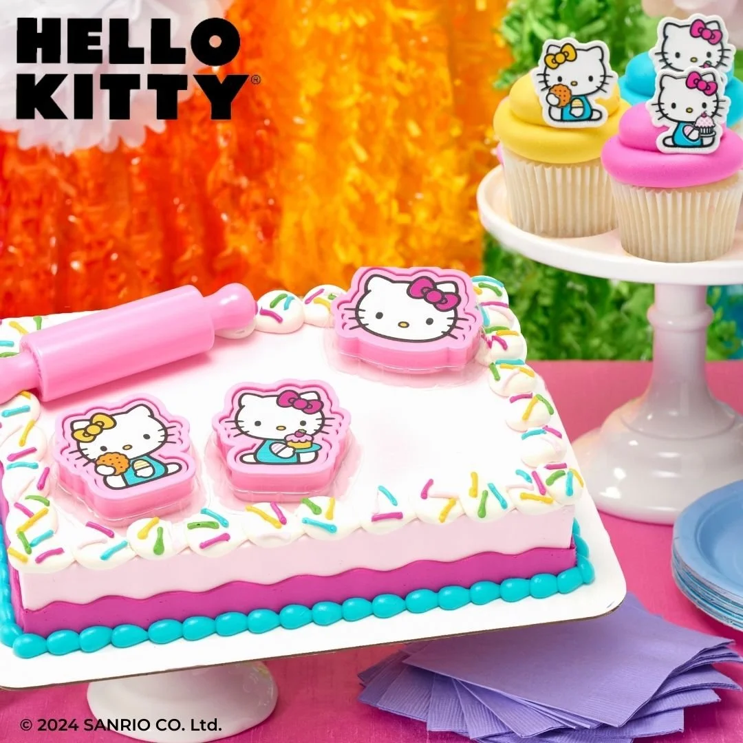 Hello Kitty themed birthday cake with colorful sprinkles and toppers, cupcakes with Hello Kitty toppers, pink tablecloth, purple napkins, and empty cake plates.