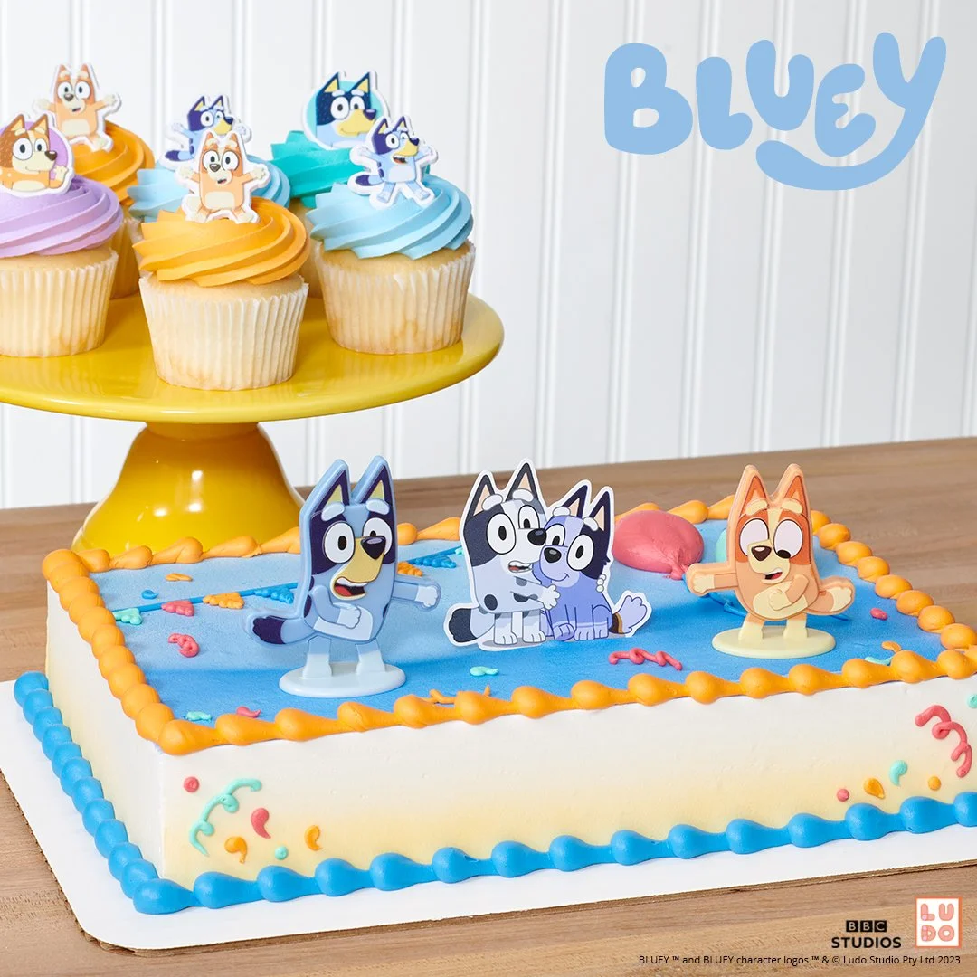 A birthday cake decorated with characters from Bluey and her family, with shaped cupcake rings on a yellow cake stand in the background.