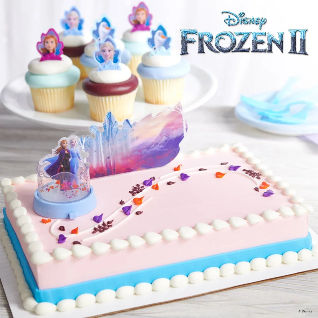 A Frozen 2-themed birthday cake with a pink top layer decorated with edible flower petals, a blue bottom border, and an ice castle topper, accompanied by cupcakes topped with Frozen characters rings.