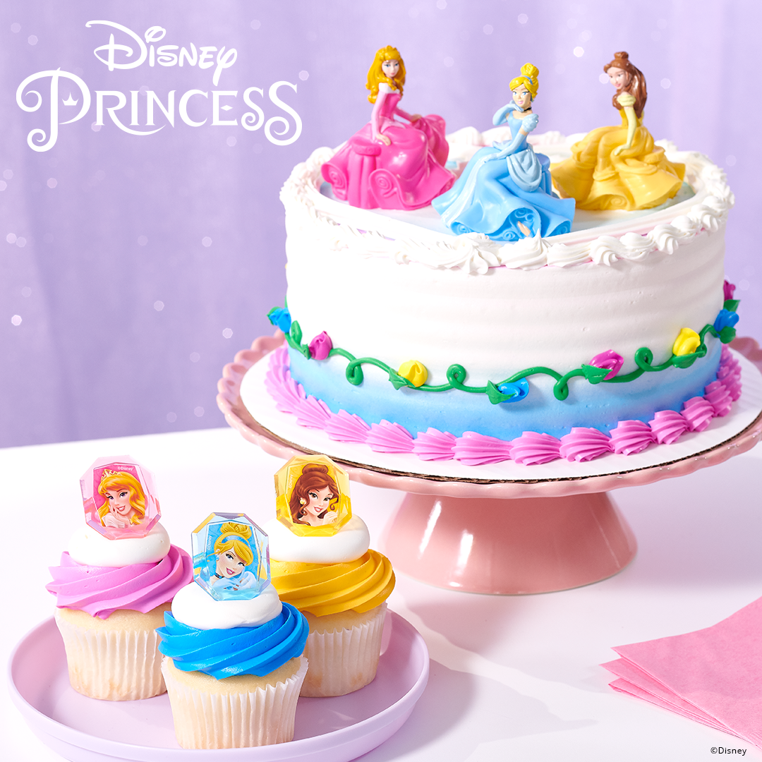A Disney princess-themed birthday cake decorated with figurines of Princess Aurora, Cinderella, and Belle, topped with white frosting and colorful decorations. In front of the cake, there are three cupcakes, each topped with a cupcake rings.