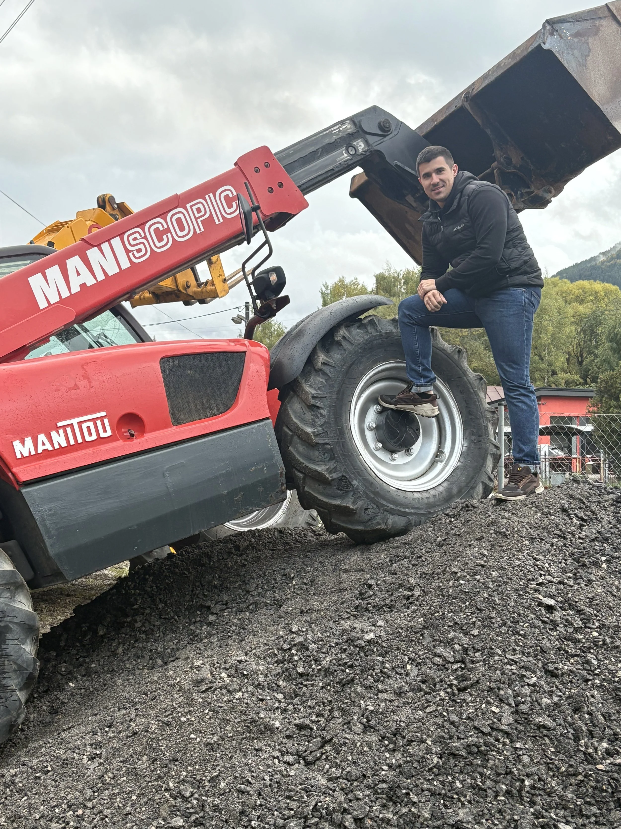 A man in black jacket and jeans is sitting on the large rear tire of a red Manitou telescopic forklift, which is positioned on a gravel surface outdoors with a cloudy sky and trees in the background.