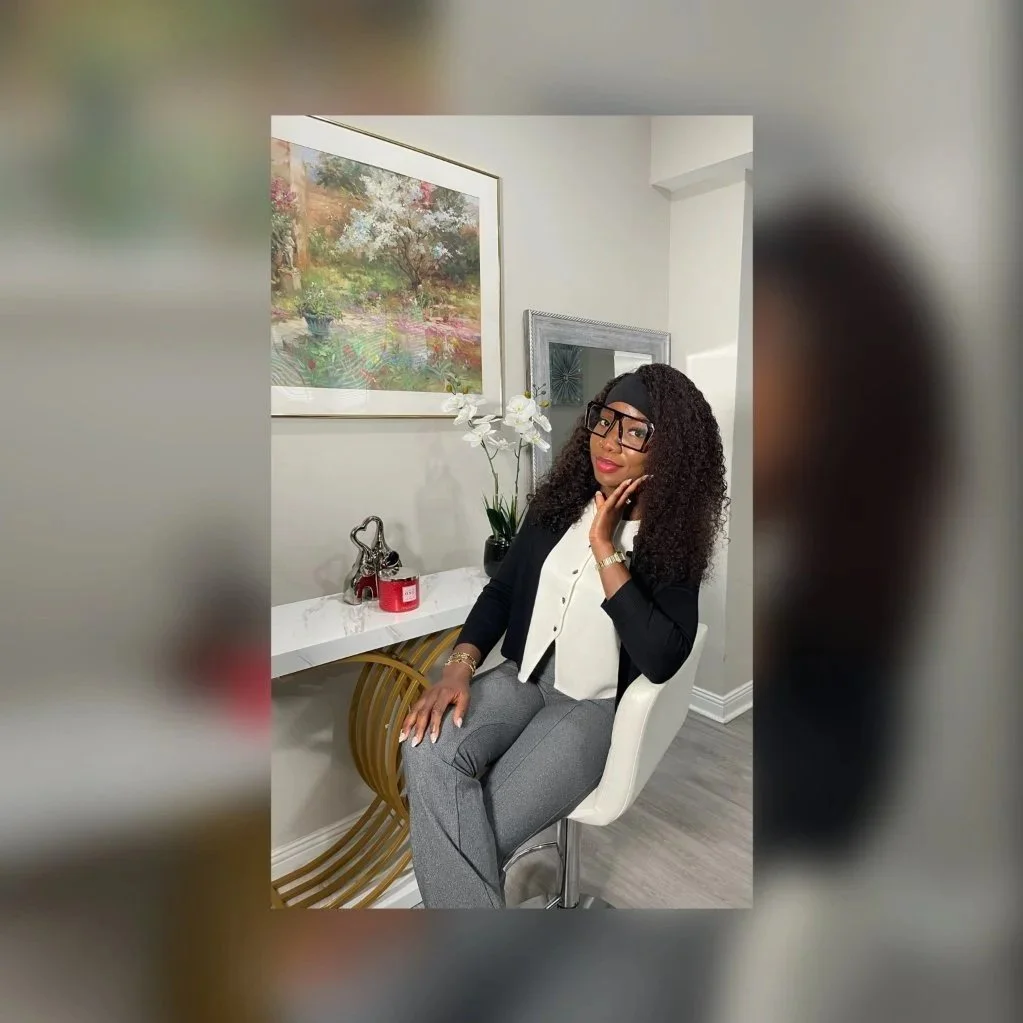 Woman with curly hair and glasses sitting on a white chair in interior room, wearing a black blazer, white blouse, and gray pants, posing with her hand on her face, with a floral painting, mirror, and decorative items on the white table behind her.