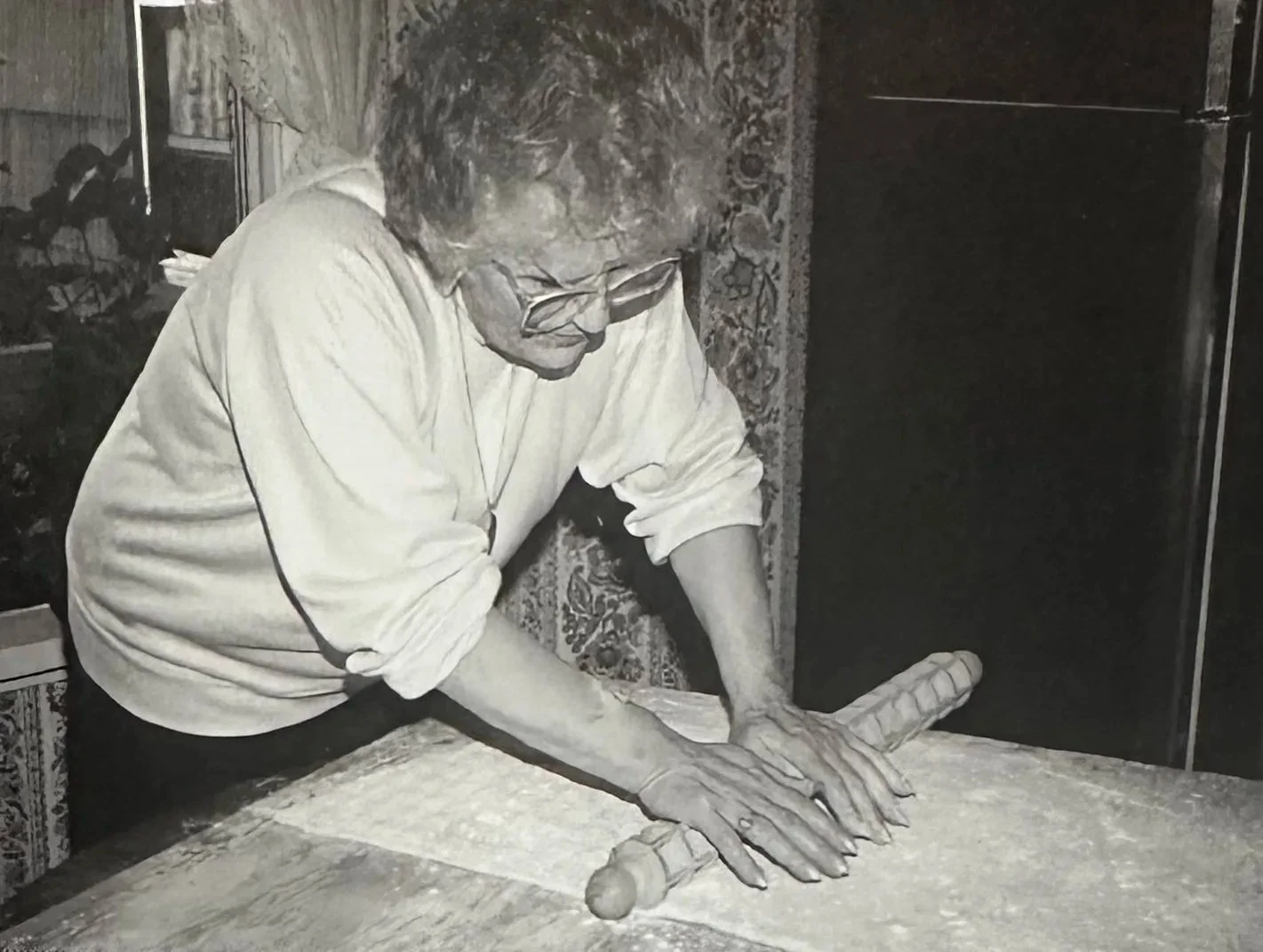 An older man with glasses, wearing a long-sleeved shirt, is rolling out dough with a rolling pin on a table.
