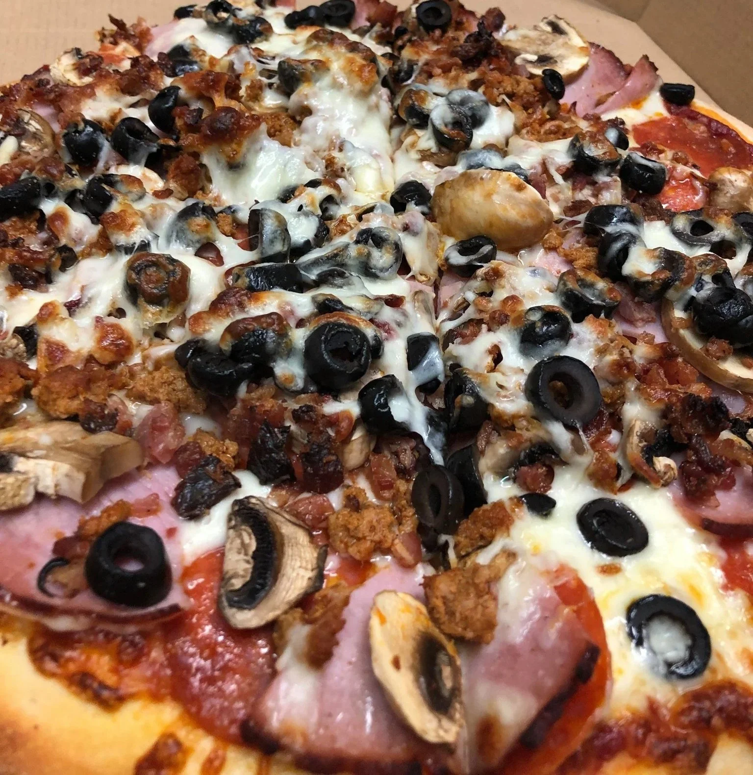 Close-up of a pizza with black olives, mushrooms, pepperoni, ham, sausage, bacon, and melted cheese.