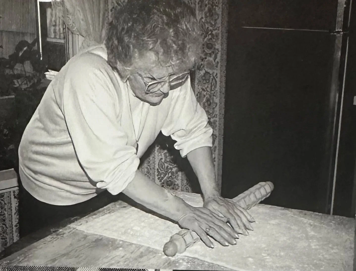 An elderly person with glasses and a white shirt rolling out dough with a rolling pin on a floured surface.
