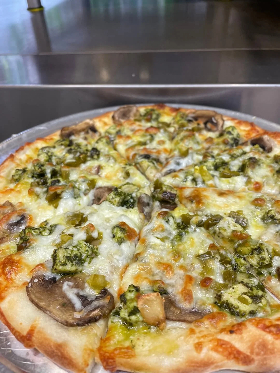 Close-up of a freshly baked vegetable pizza with cheese, mushrooms, green peppers, and possibly broccoli on a metal tray.
