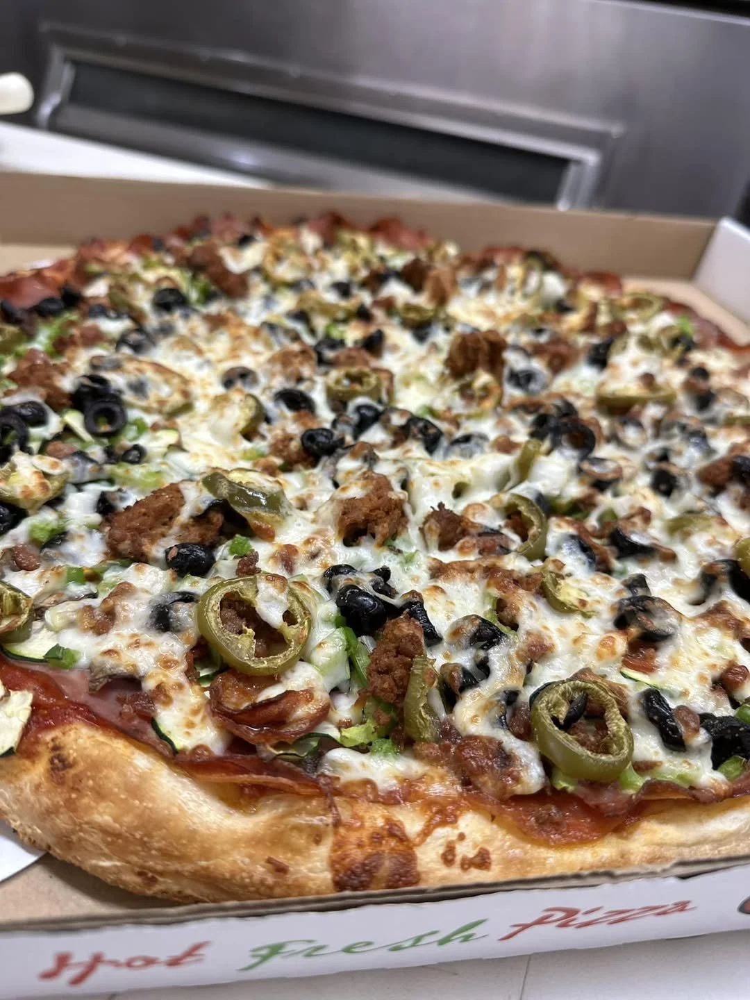 Close-up of a freshly baked pizza topped with black olives, green peppers, sausage, and cheese in a cardboard box.