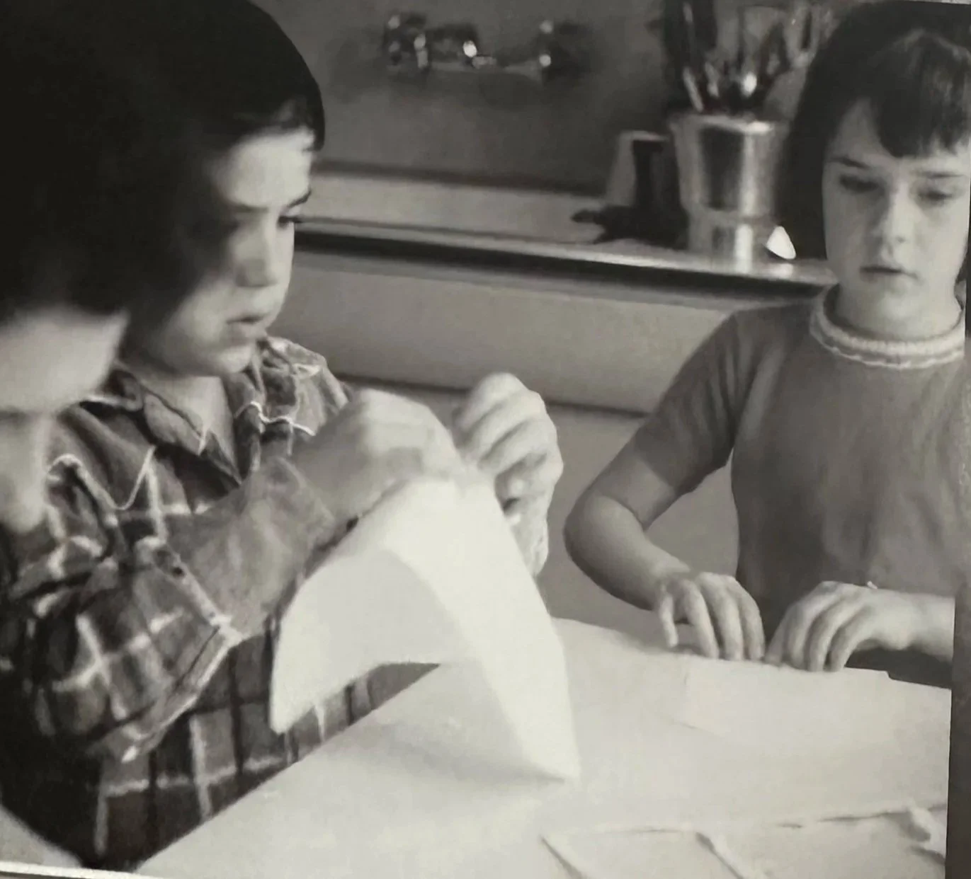 Three children sitting at a table, with two of them focused on a paper shaped like a fish, in black and white.