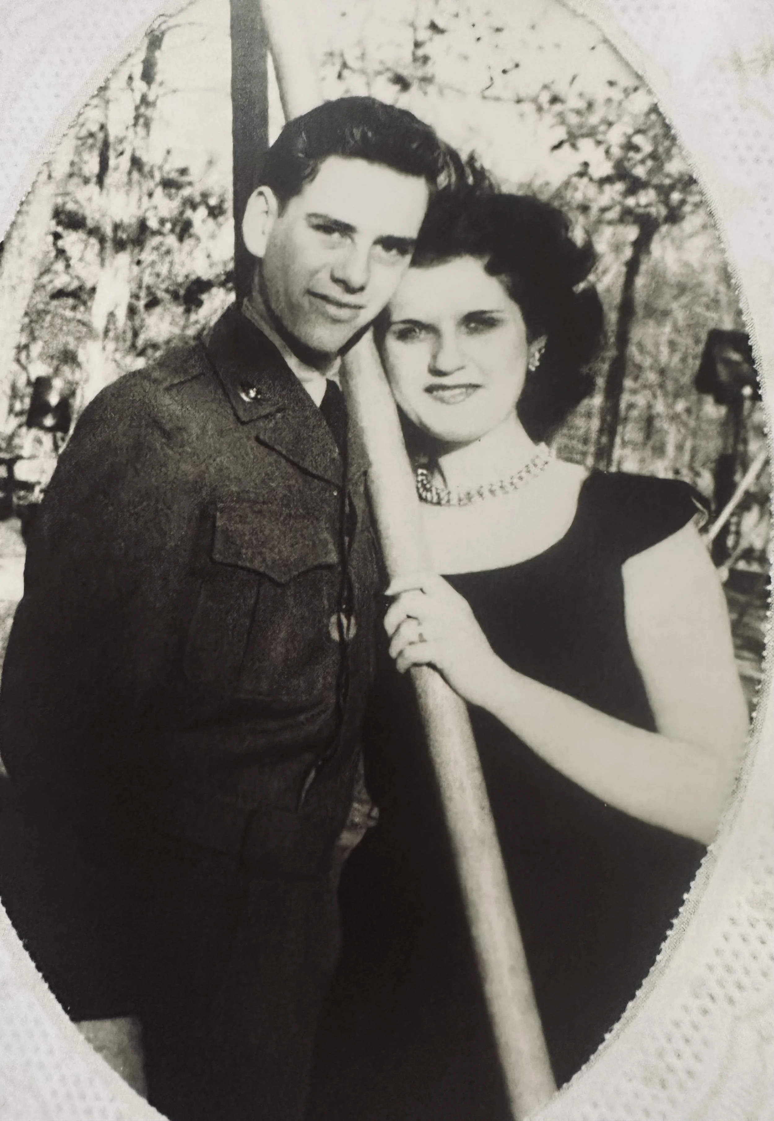 A black-and-white photograph of a man in a military uniform and a woman in a dark dress, standing outdoors, with trees and a flag in the background. The woman is holding a flagpole.