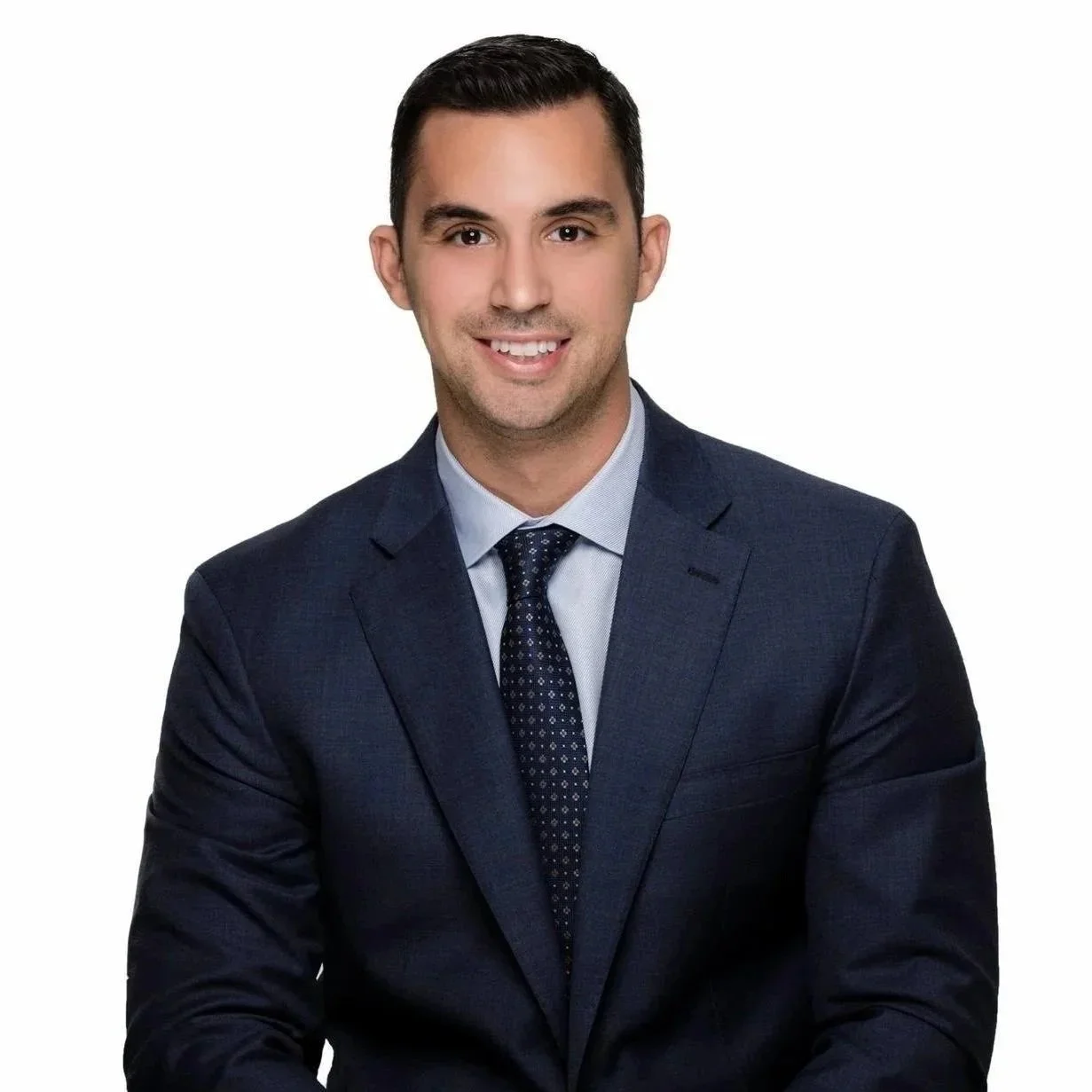 A professional young man with dark hair in a navy blue suit, light blue shirt, and patterned tie, smiling, against a white background.