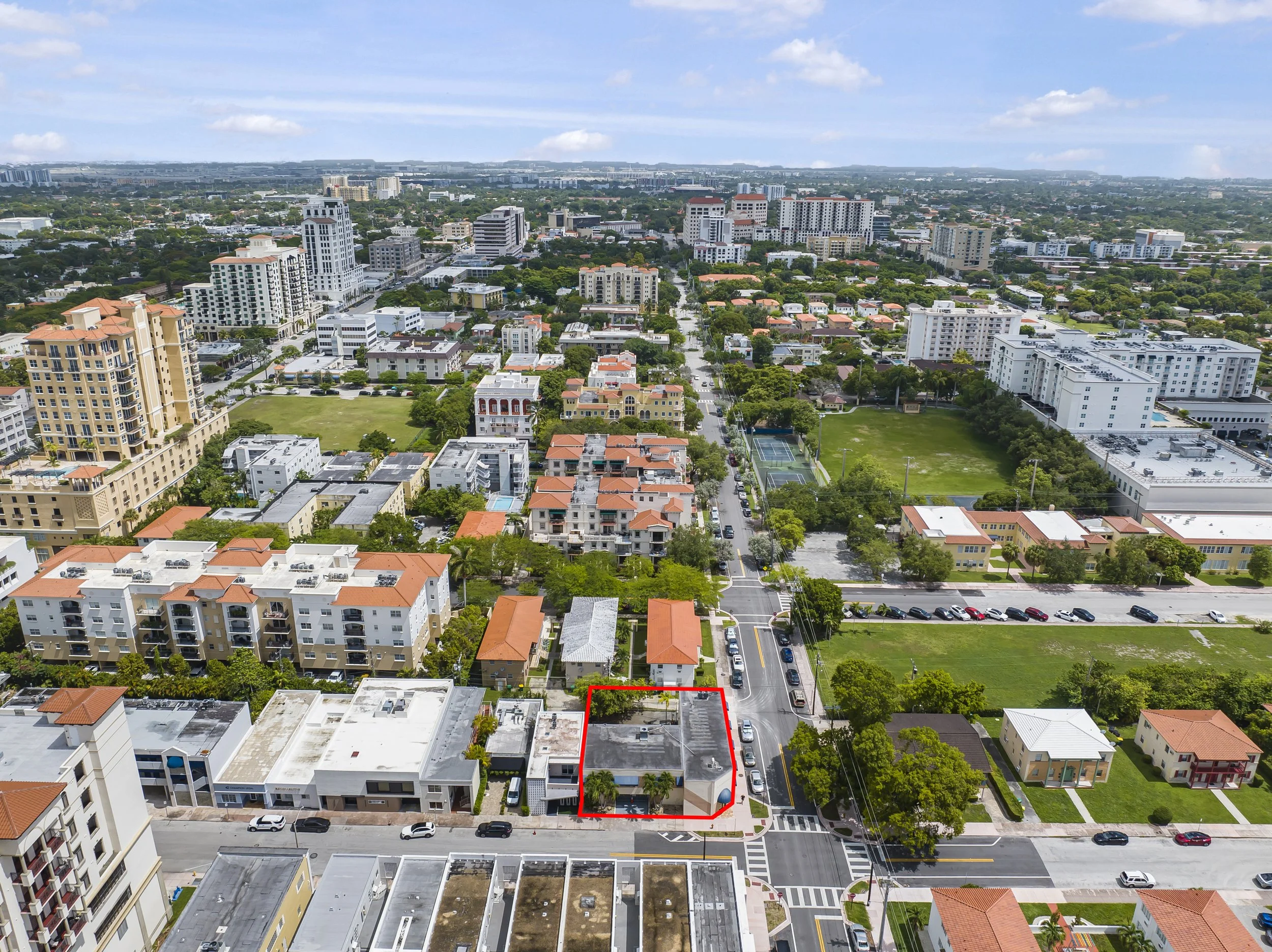 Aerial view of a cityscape with various mid-rise and high-rise buildings, green parks, and streets, with a red outline around a small building at the intersection of two streets.