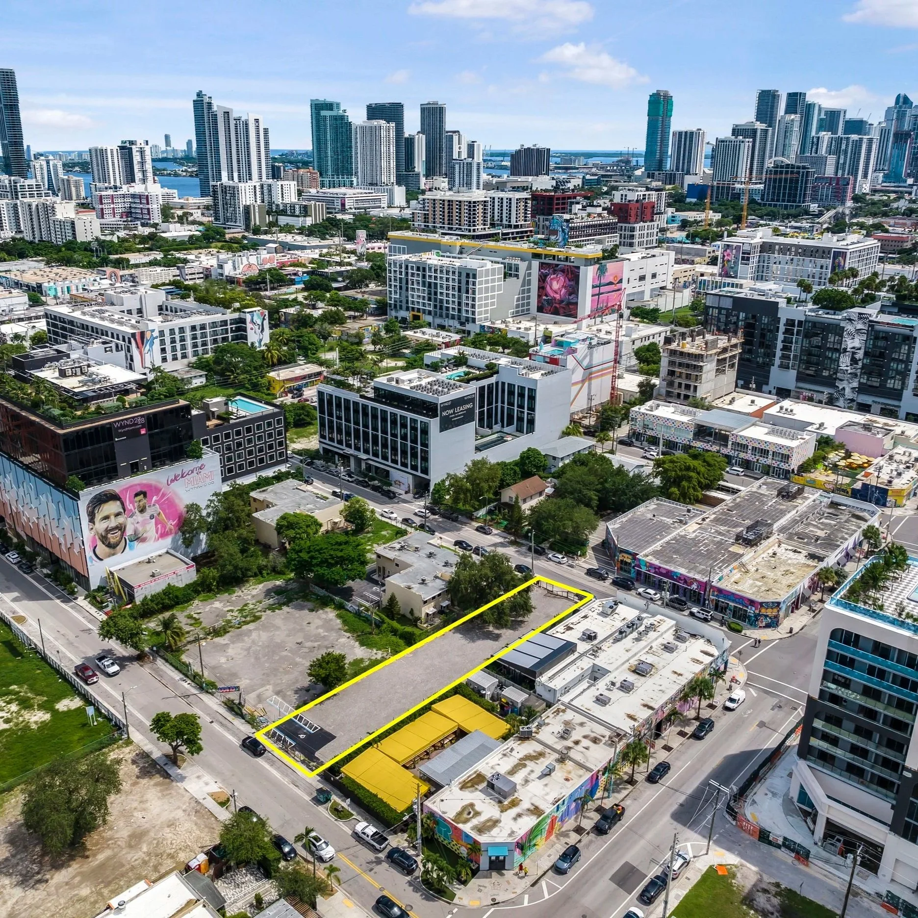 Aerial view of downtown Miami, Florida with tall skyscrapers, colorful murals on buildings, and a mix of commercial and residential structures. A parking lot and a small building with bright yellow awnings are in the foreground.
