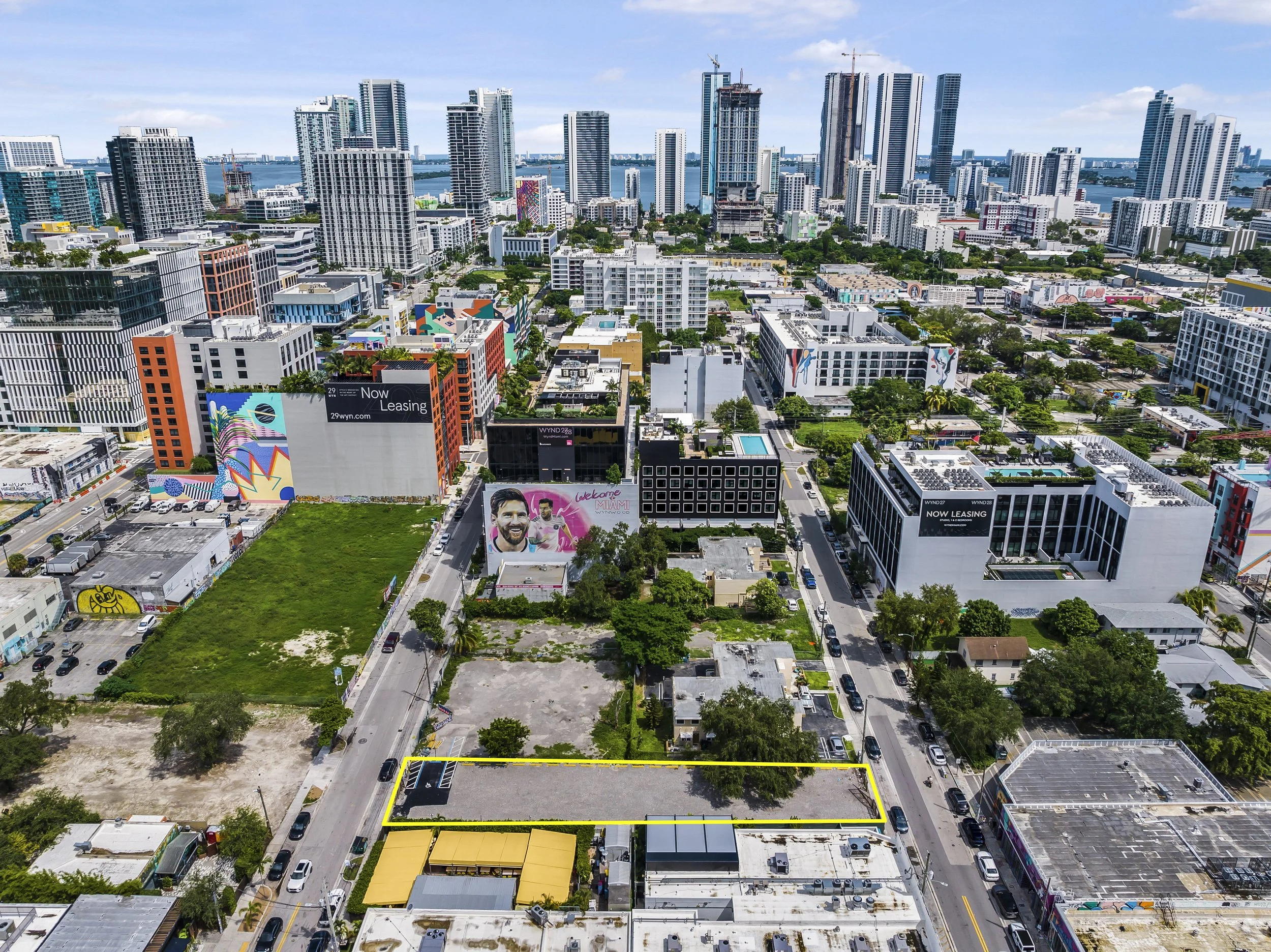 Aerial view of Miami cityscape with high-rise buildings, colorful murals, and a vacant lot outlined in yellow, under a partly cloudy sky.