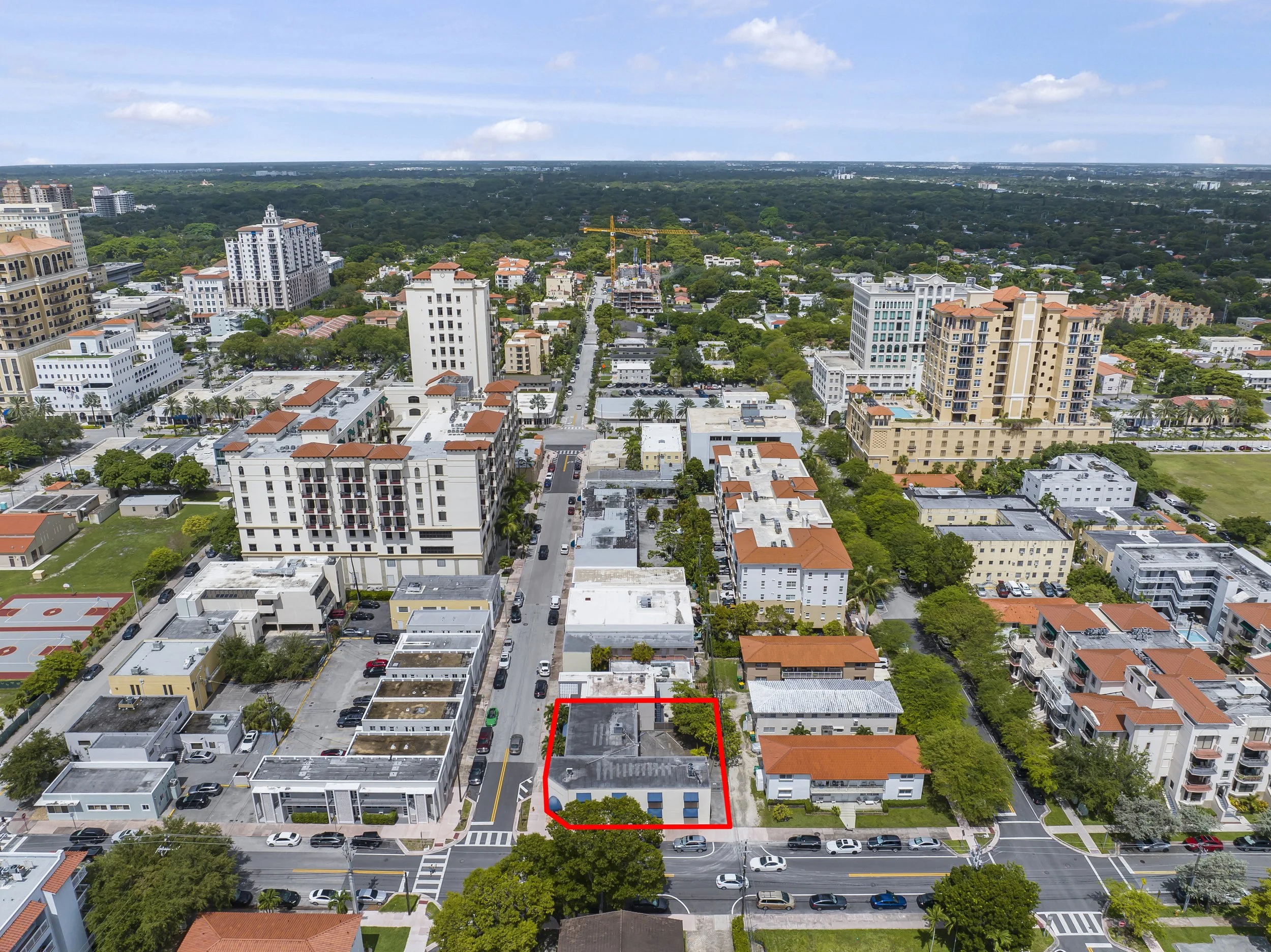 Aerial view of a city with residential and commercial buildings, streets lined with cars, and a outlined property at the center with red border.