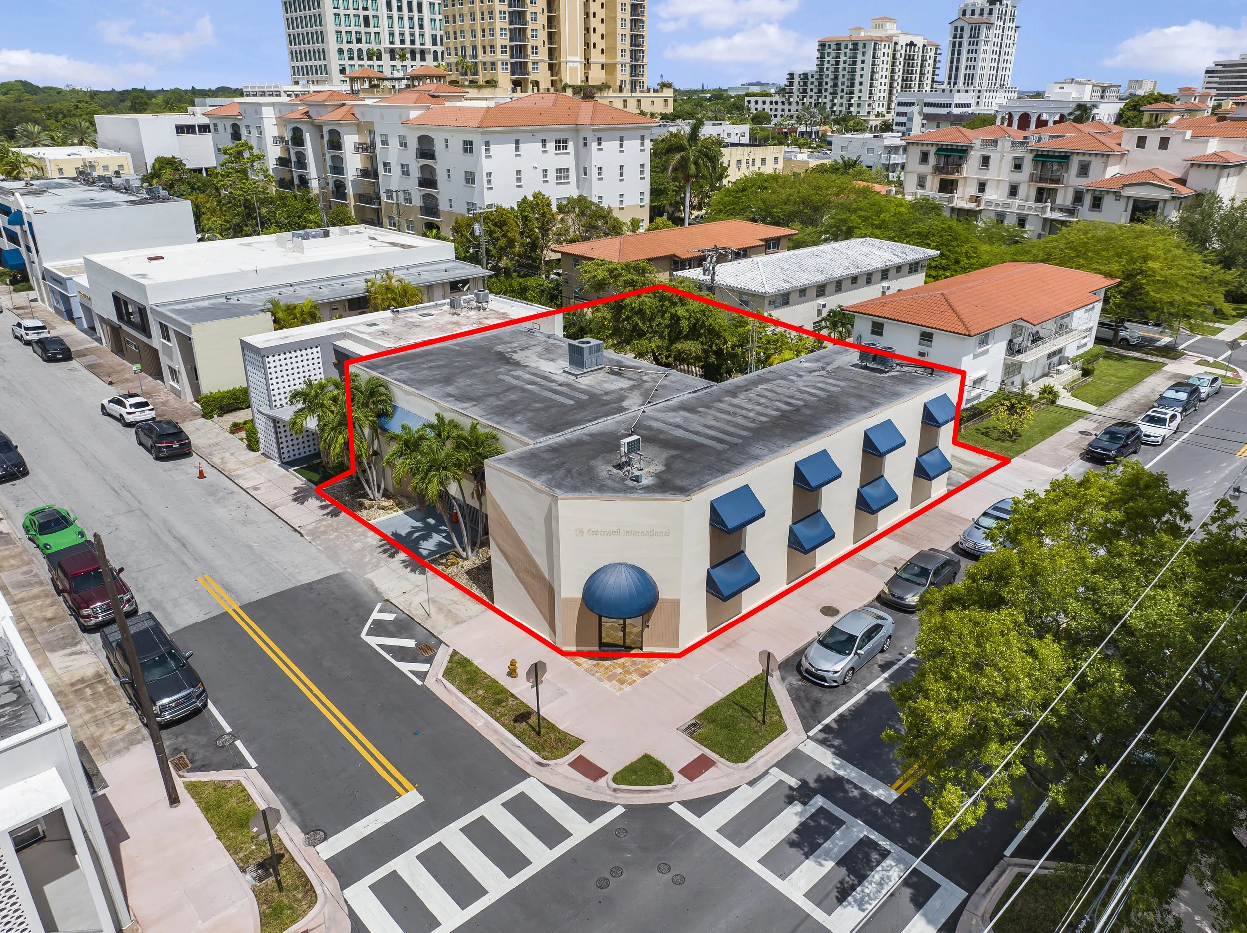 Aerial view of a commercial building on a city street with parking spaces, trees, and neighboring residential and commercial buildings, with a red outline around the building.