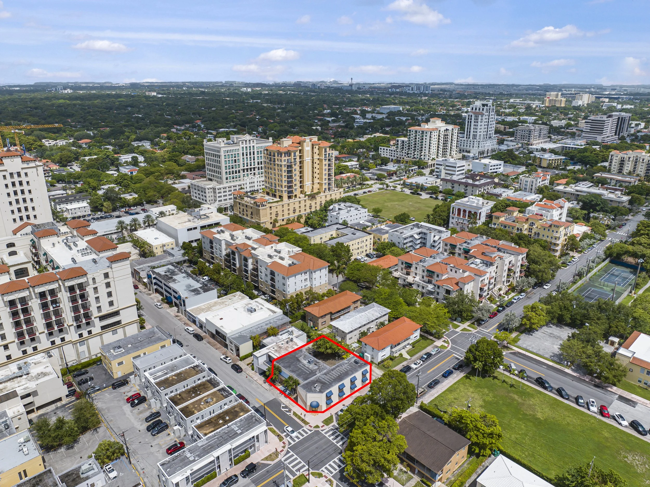 Aerial view of a city landscape with various residential and commercial buildings, roads, and green spaces, including tennis courts and a park.