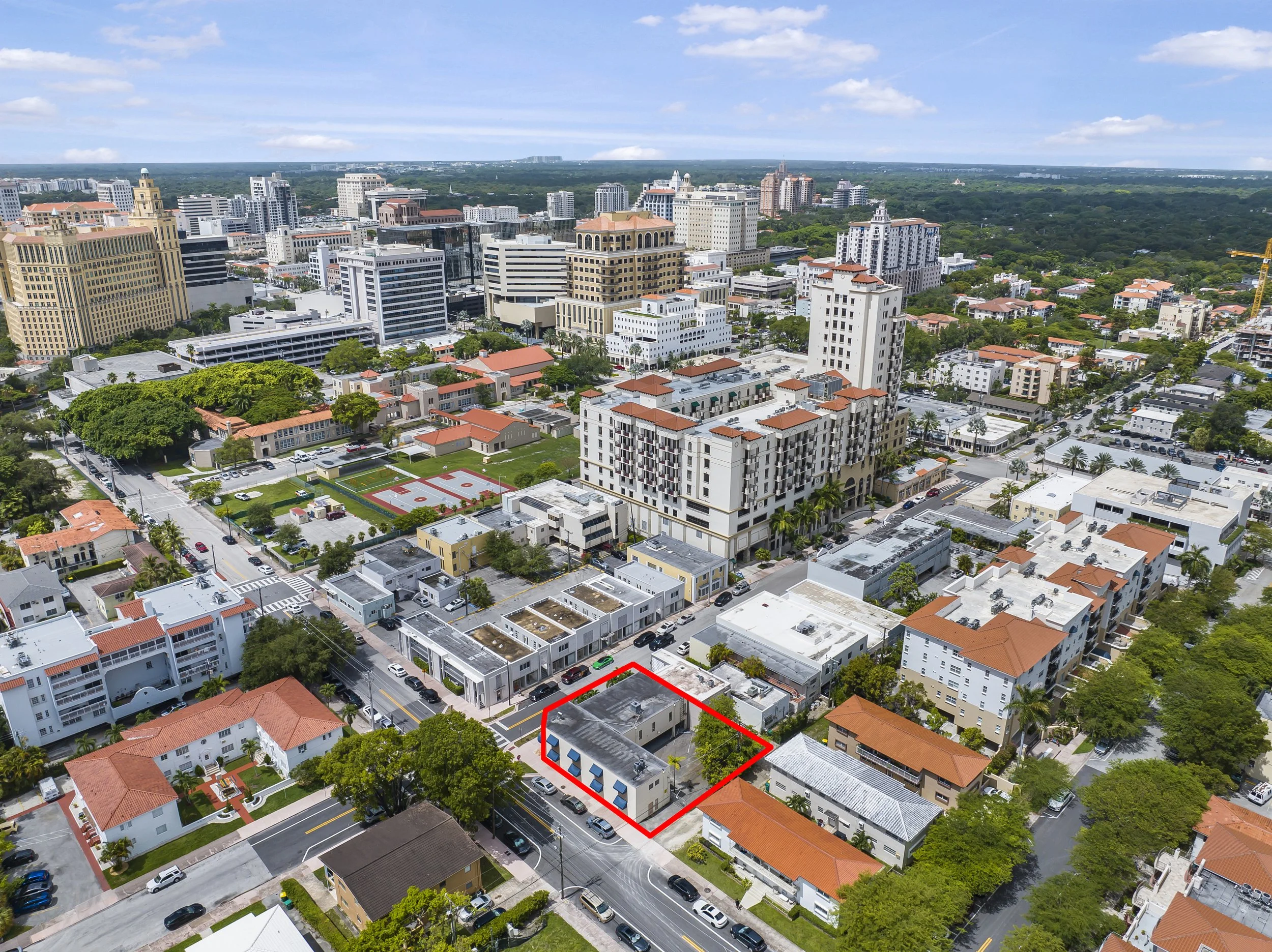 Aerial view of a city with a mix of modern high-rise buildings and smaller residential houses. One building is outlined in red, located near a main road with trees and parking areas nearby.