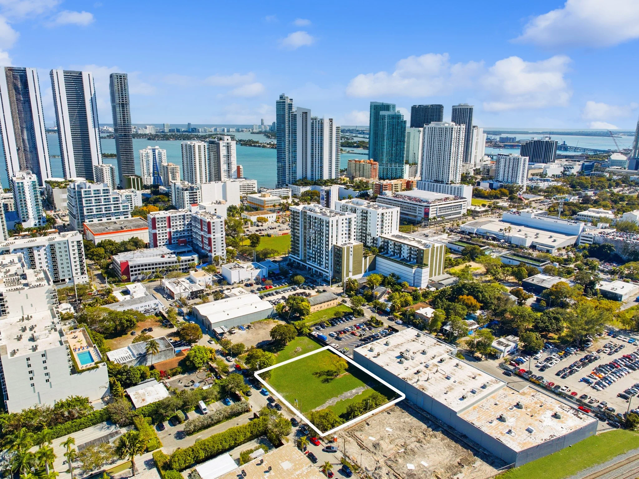 Aerial view of a cityscape with high-rise buildings, water bodies, and residential and commercial areas, featuring a vacant grassy lot outlined in white in the foreground.