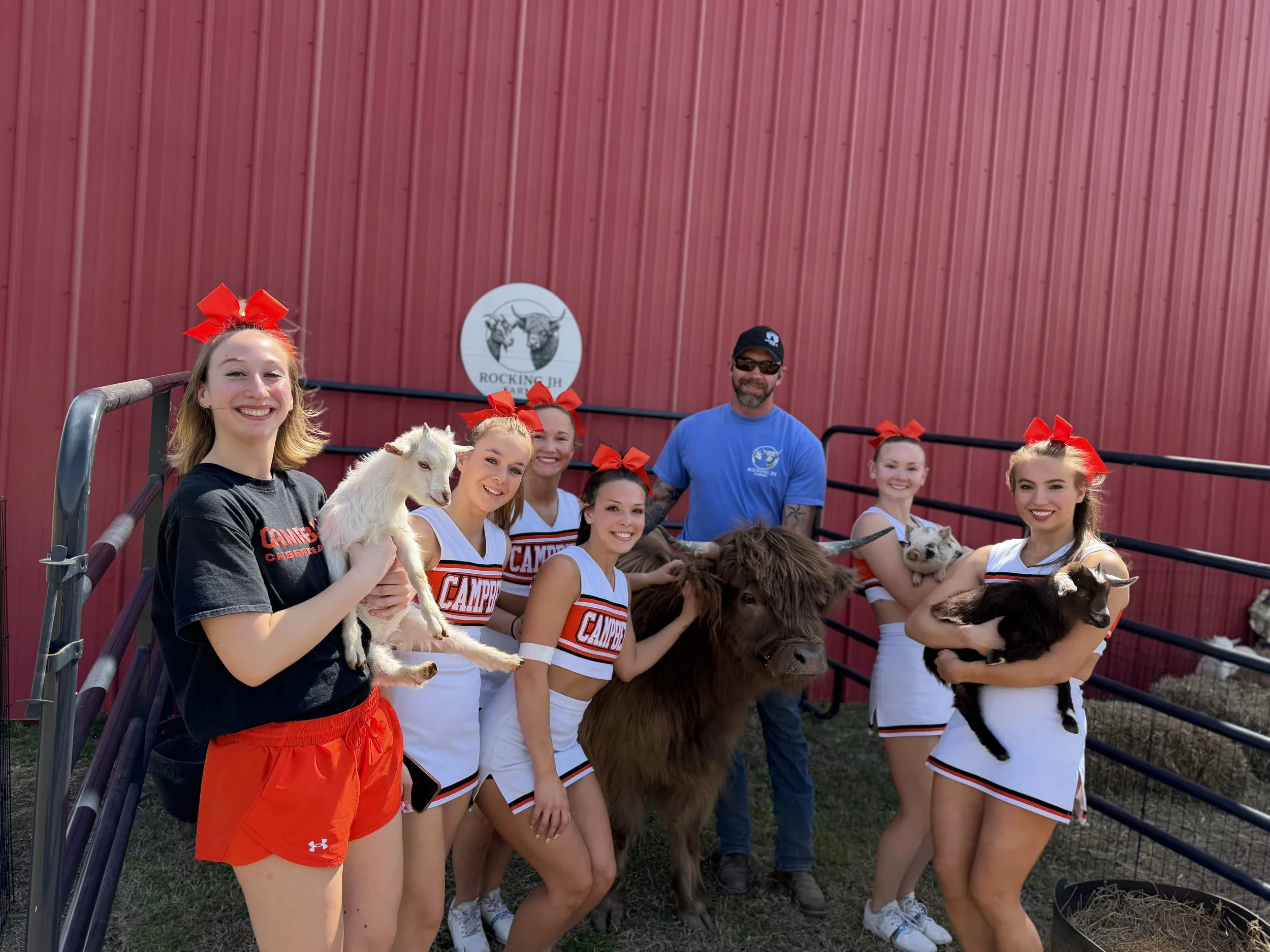 Group of cheerleaders and a man holding goats at a farm or ranch, with a red barn in the background.