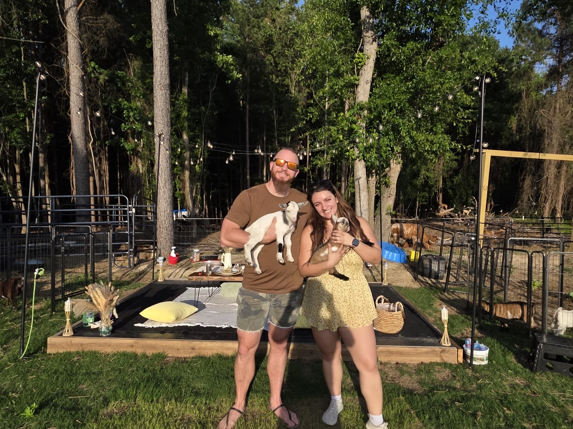 A man and woman standing outdoors with a black and white goat, smiling, in front of a decorated table and string lights among trees.