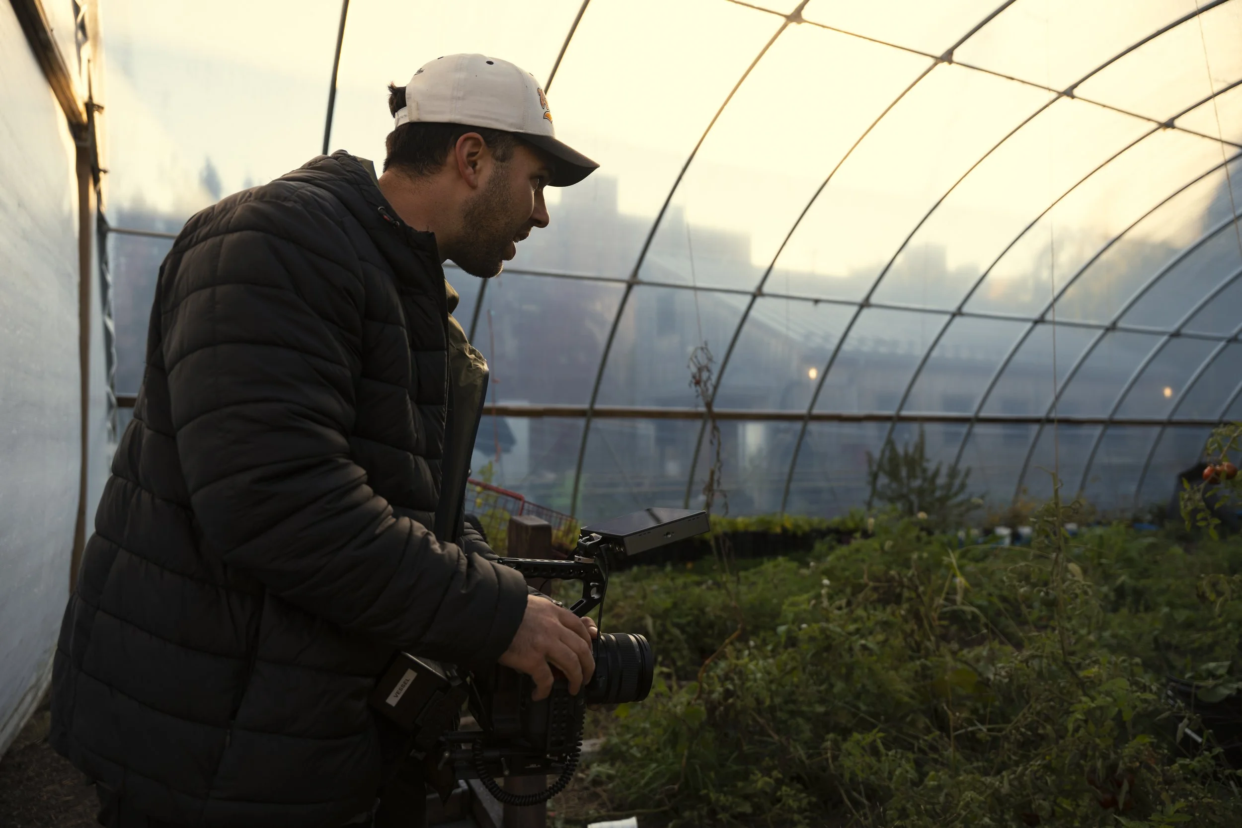 A man wearing a white cap and black puffer jacket working in a greenhouse, holding a camera or electronic device, with plants and a city skyline in the background at sunset.