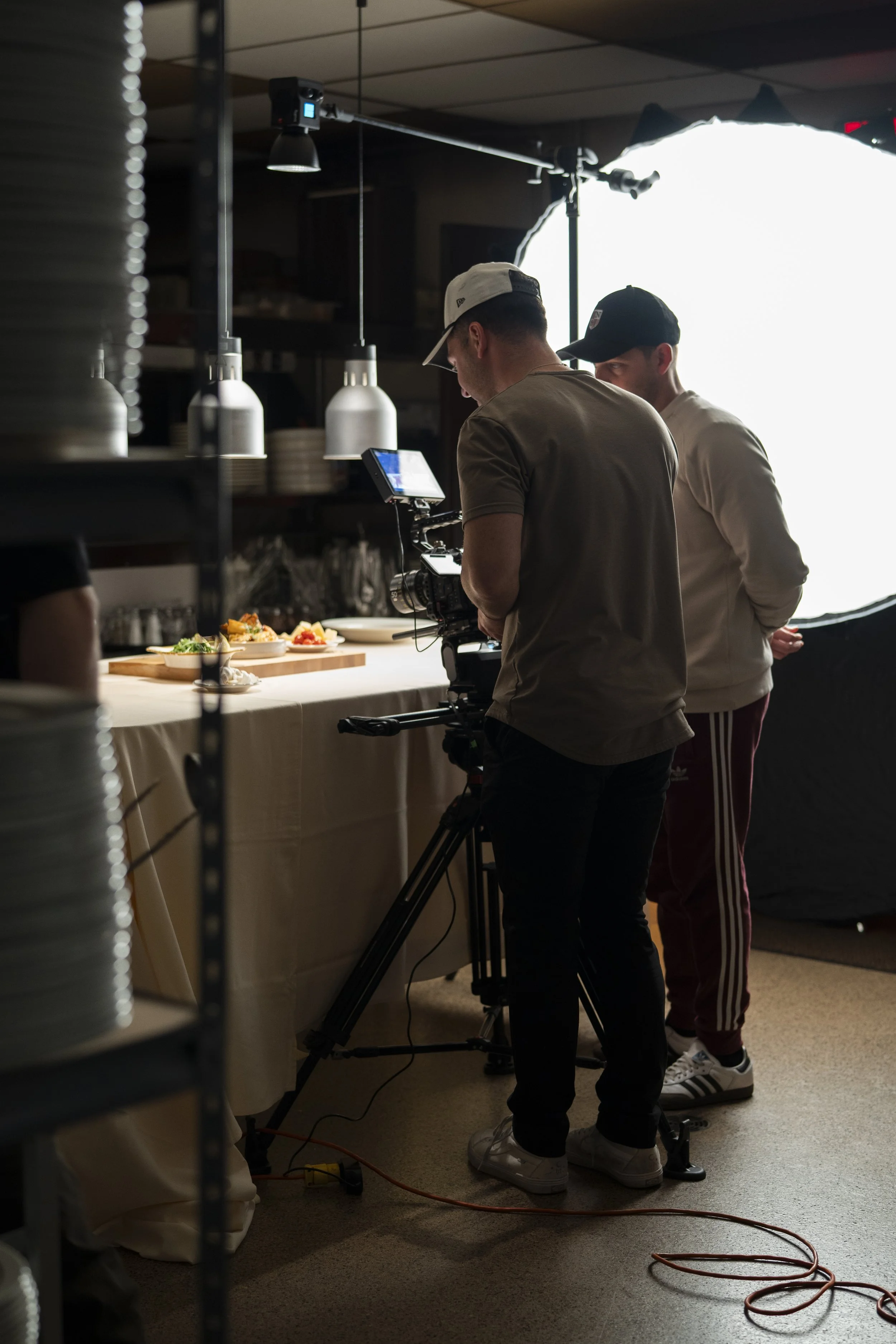 Two men are filming in a restaurant kitchen. One man operates a camera, and the other stands nearby. There are dishes of food on the counter, and lighting equipment is visible.