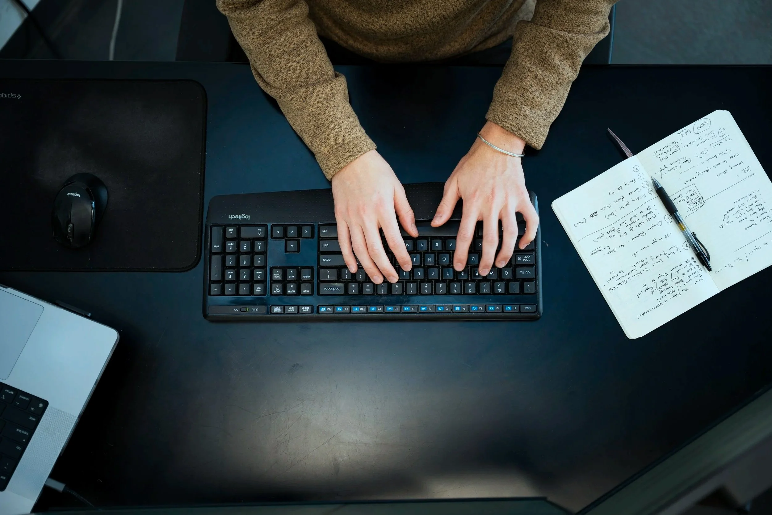 Top-down view of a person typing on a black keyboard, with a mouse, a notebook with handwritten notes, and a laptop on the desk.