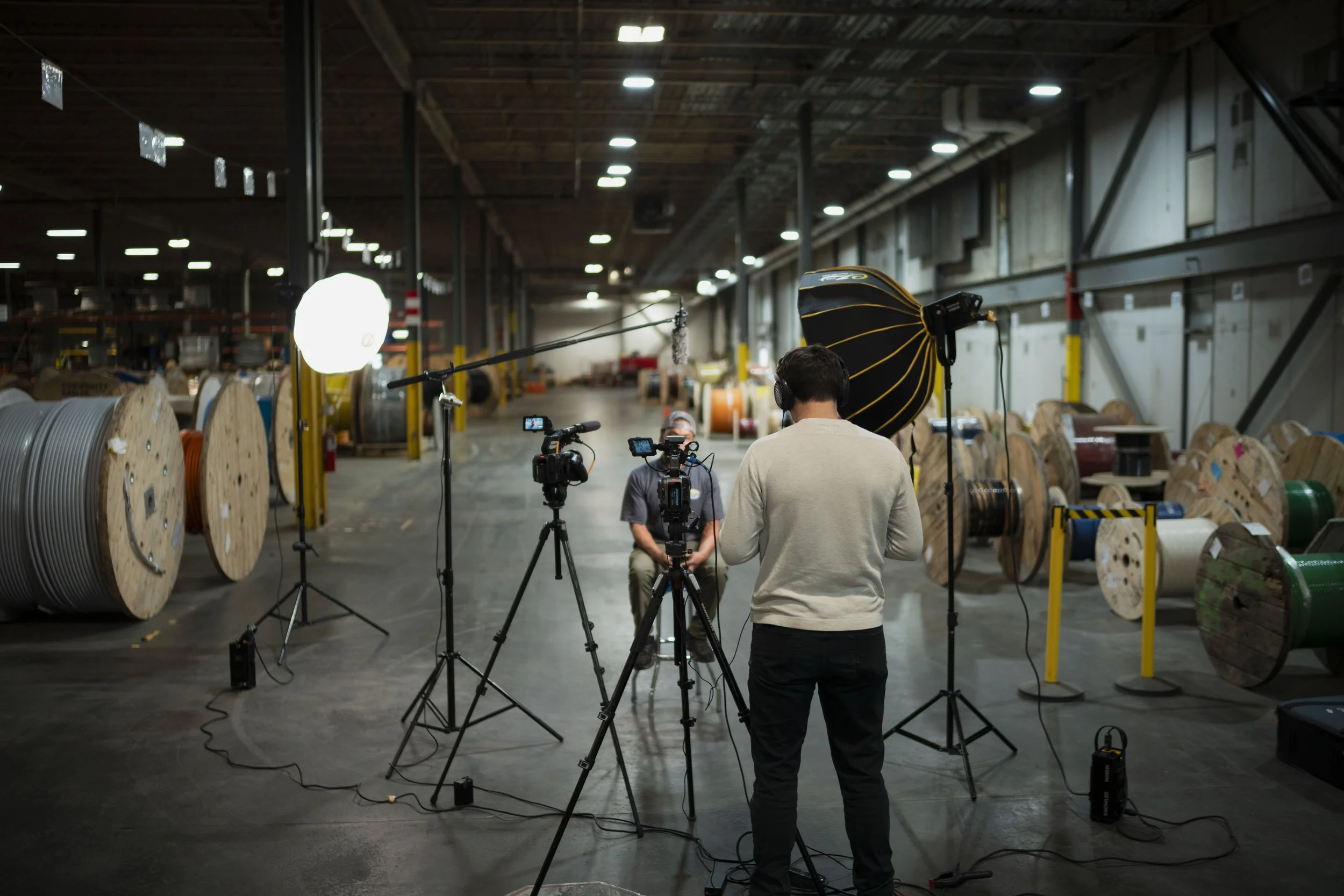 Behind-the-scenes of a video shoot in a warehouse with large spools of cable on the left and production equipment, two crew members are working with cameras and lighting.