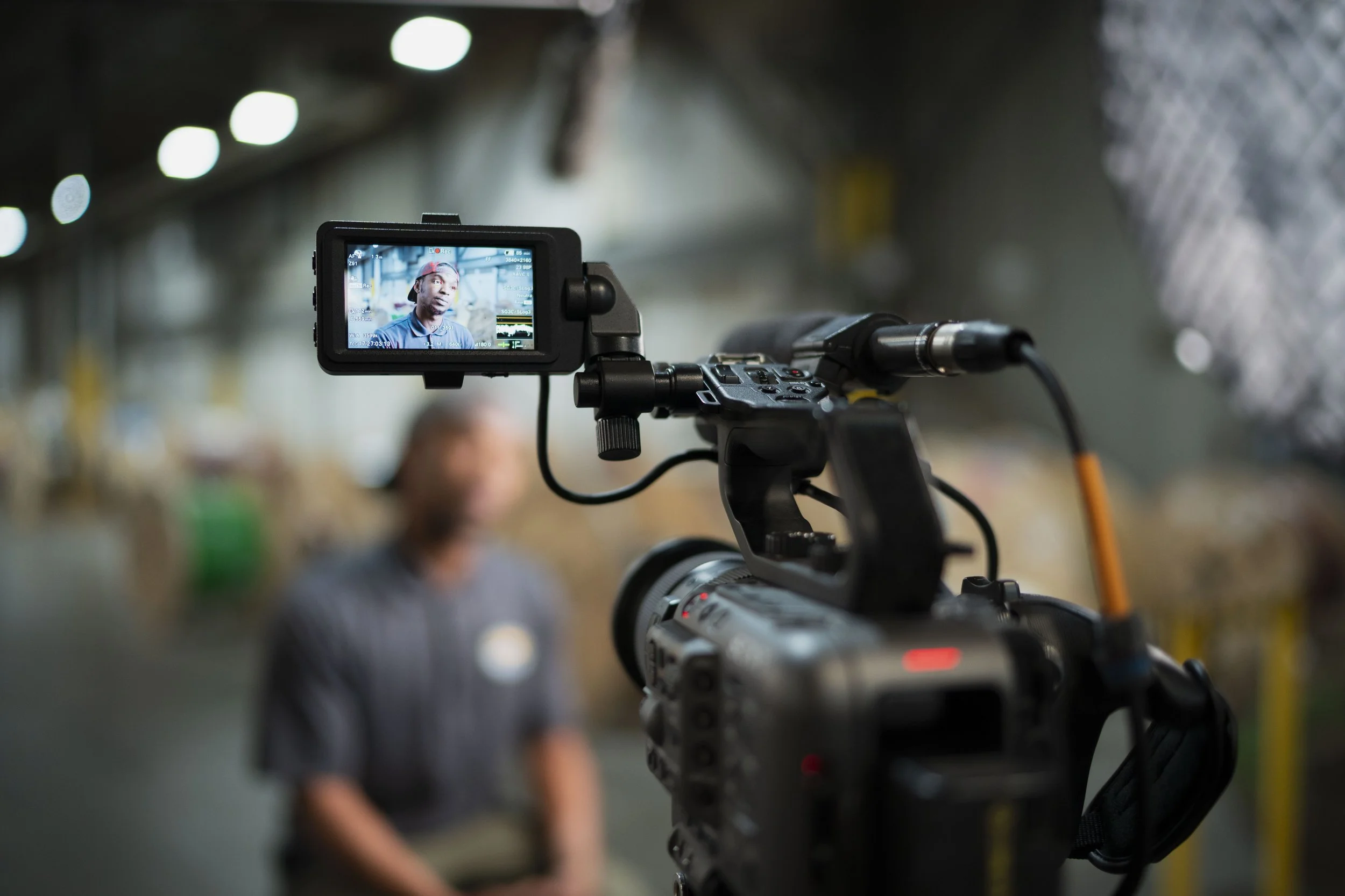 A video camera on a tripod recording a man in a gray shirt in an industrial setting.