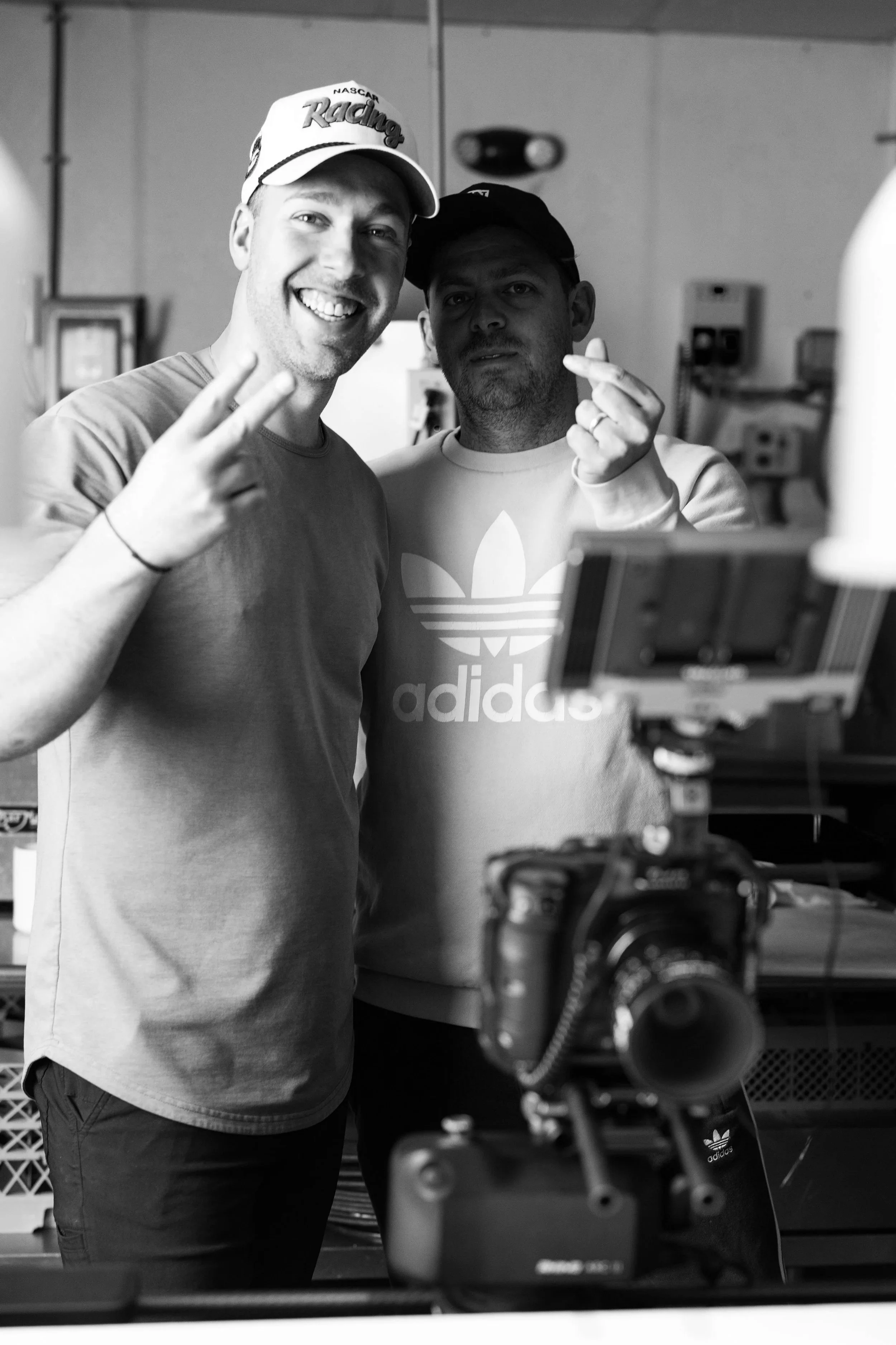 Two men taking a selfie in a room with various electronic equipment. One man is smiling widely and showing a peace sign, while the other is making a finger heart gesture. The camera is visible in the foreground.