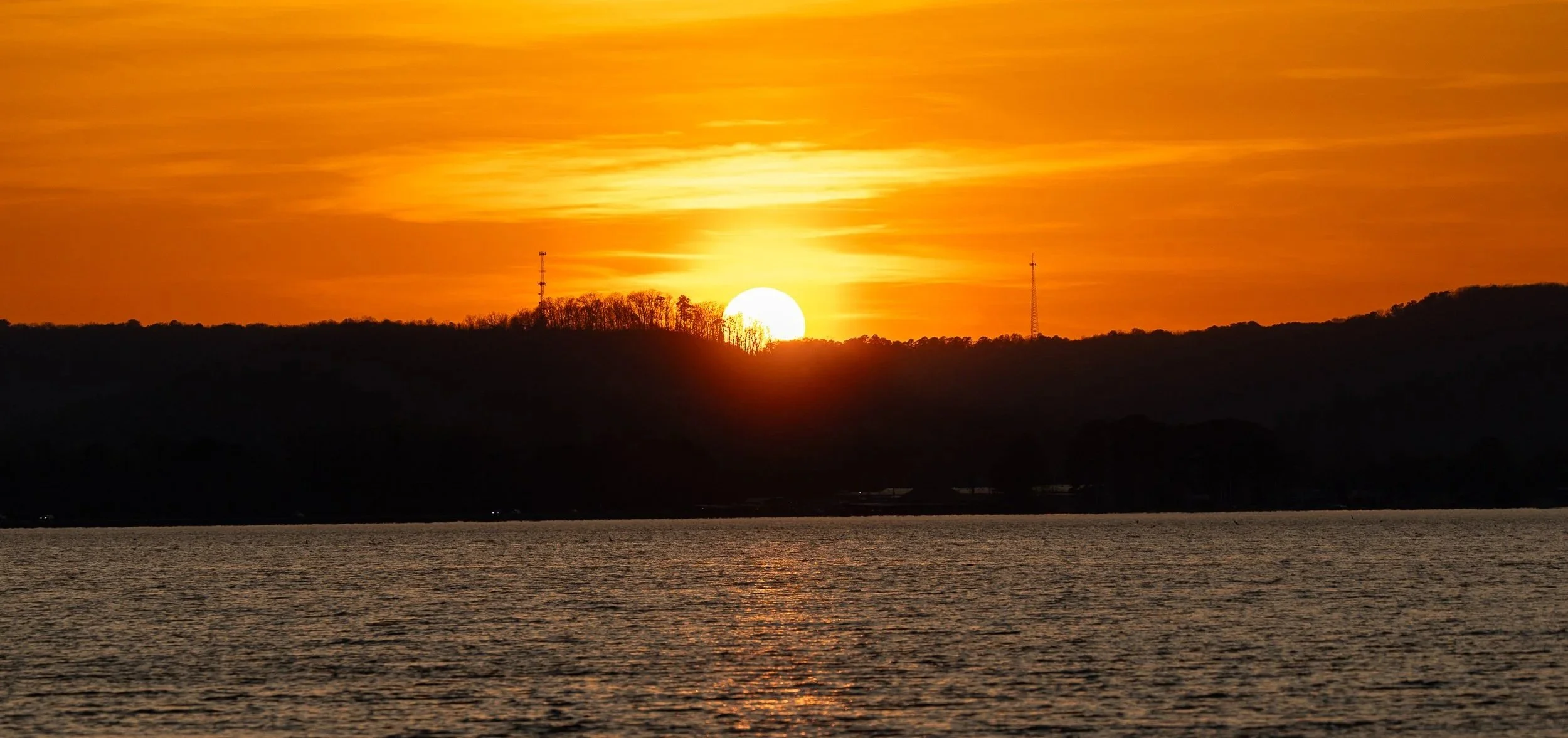 Sunset over a lake with silhouette of trees and hills in the background, and two tall communication towers on the horizon.