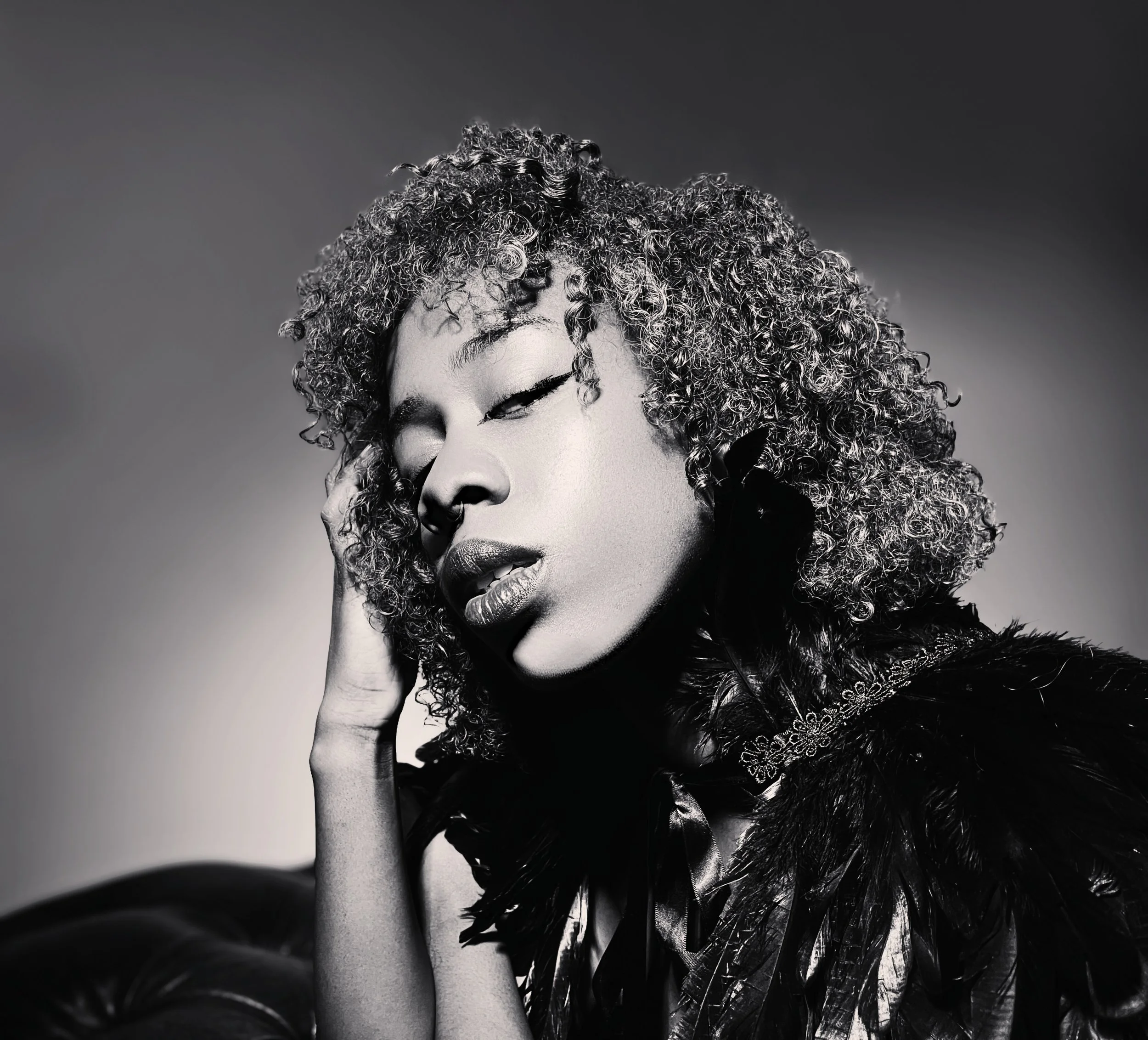 A black and white portrait of a woman with short, curly hair, closed eyes, and a hand gently touching her face, wearing a feathered garment.